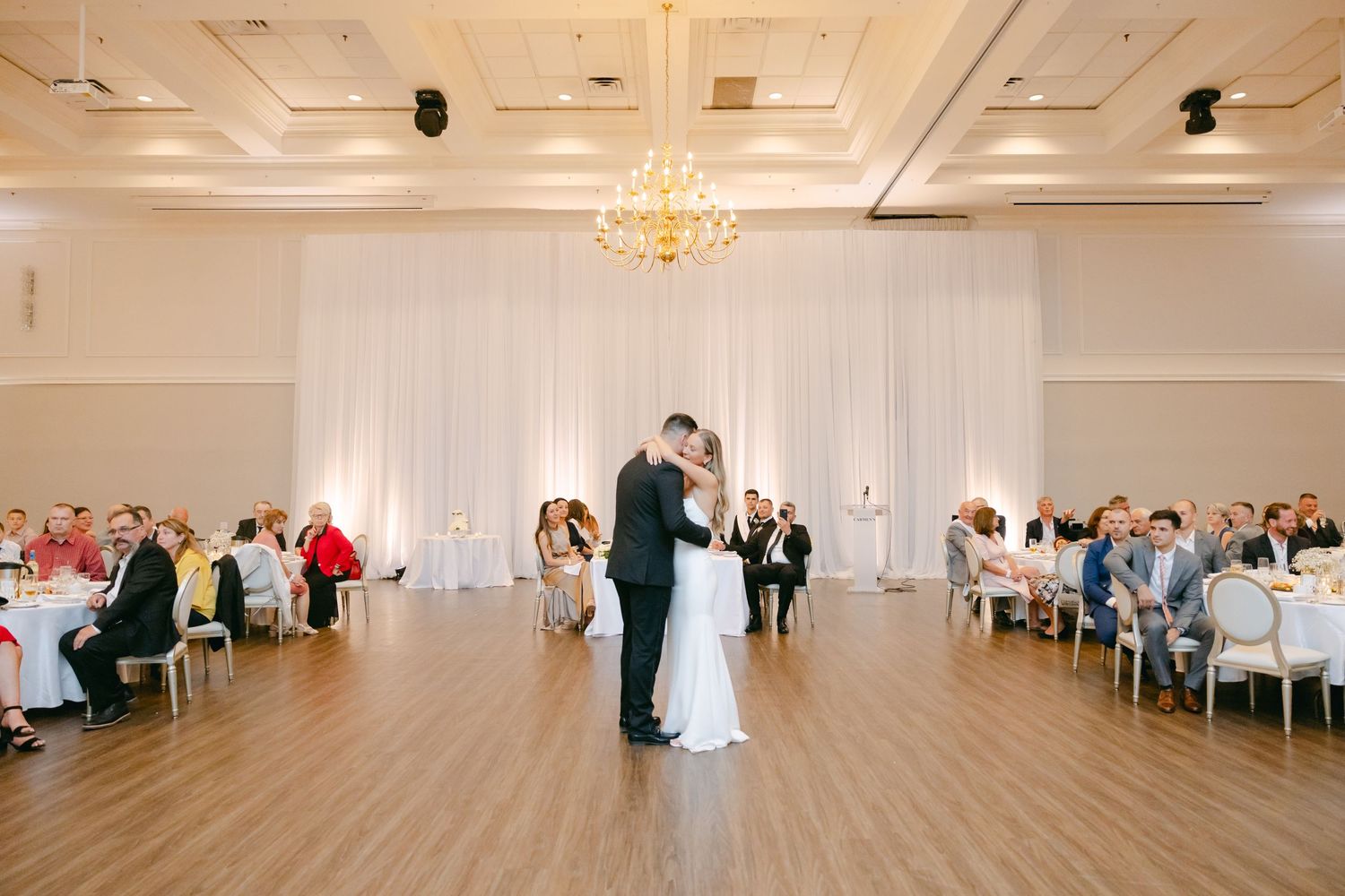 Wedding reception dance in a bright ballroom with guests seated at round tables.