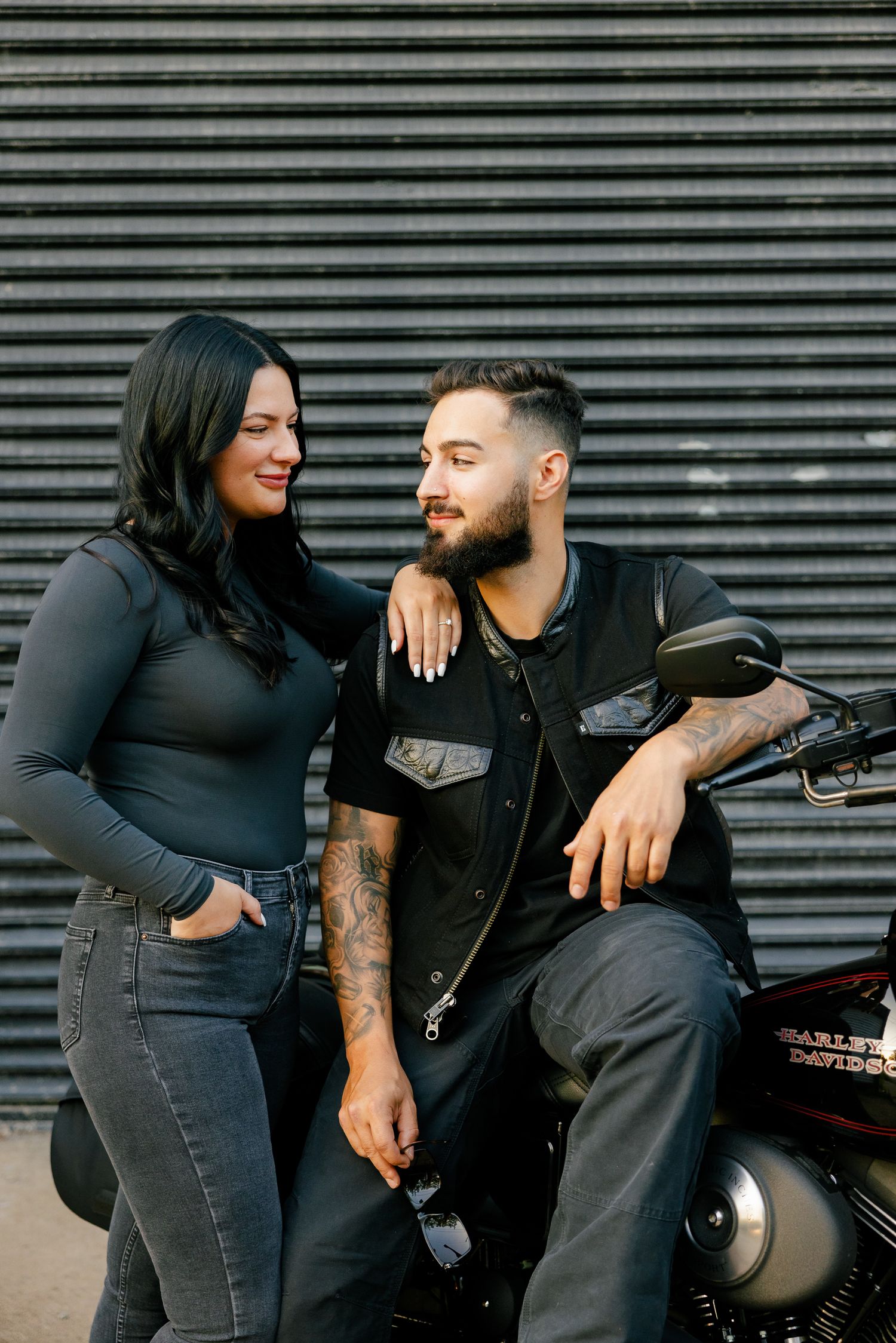 A couple shares a candid moment while posing with their black motorcycle against industrial backdrop.