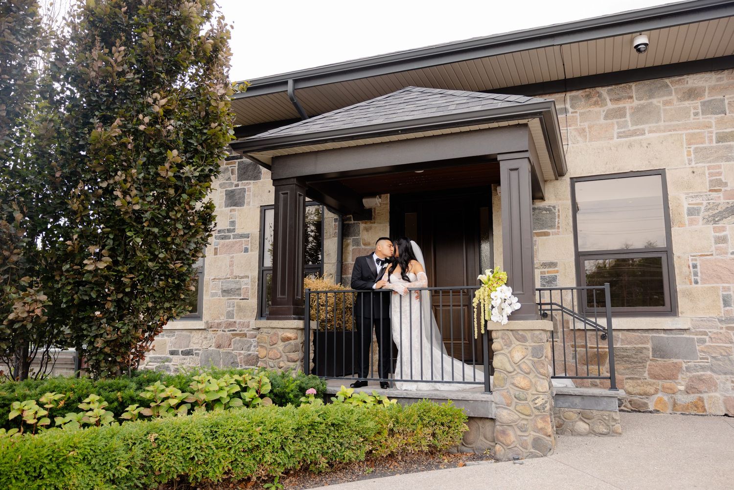Wedding couple stands in elegant stone entryway with columns and landscaping at formal venue entrance.