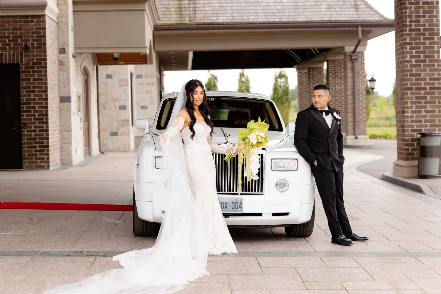 A wedding party poses together outside a brick building entrance with white columns and a white limousine visible.