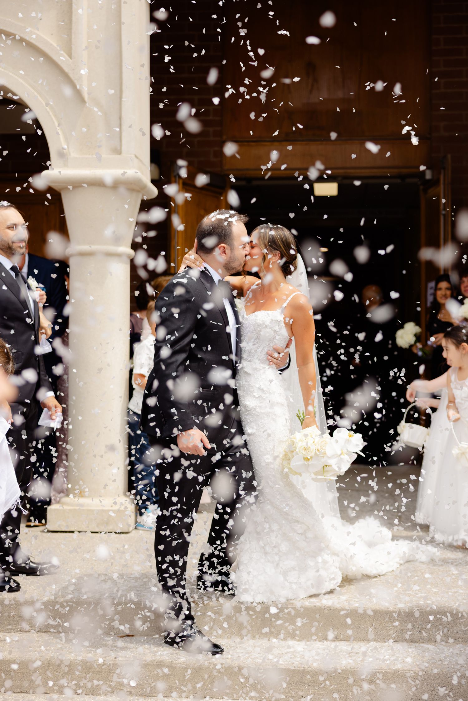 Wedding couple embraces under stone archway while guests shower them with white confetti in celebratory moment.