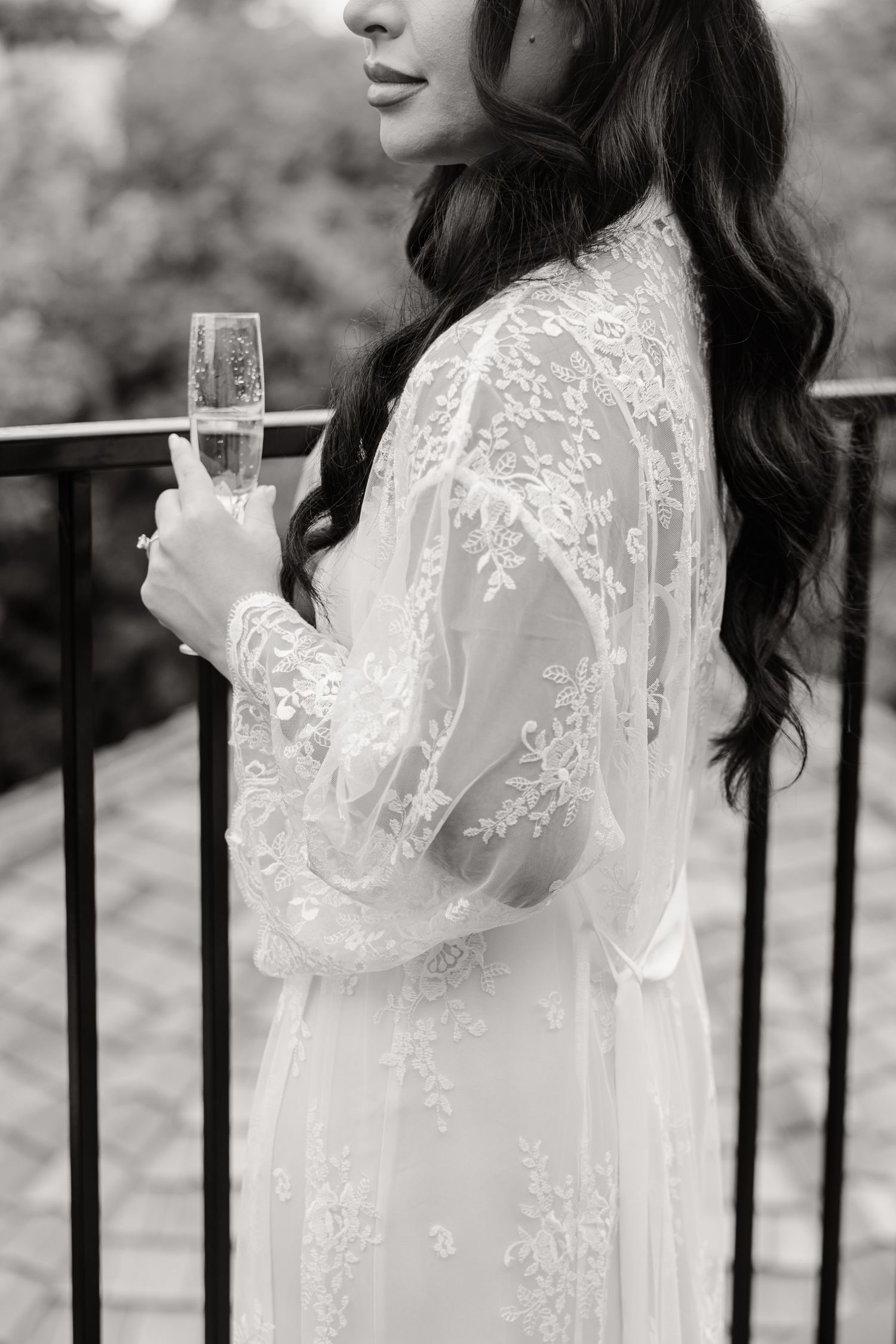 A person in a delicate white lace robe holds a glass while standing against an iron railing outdoors.