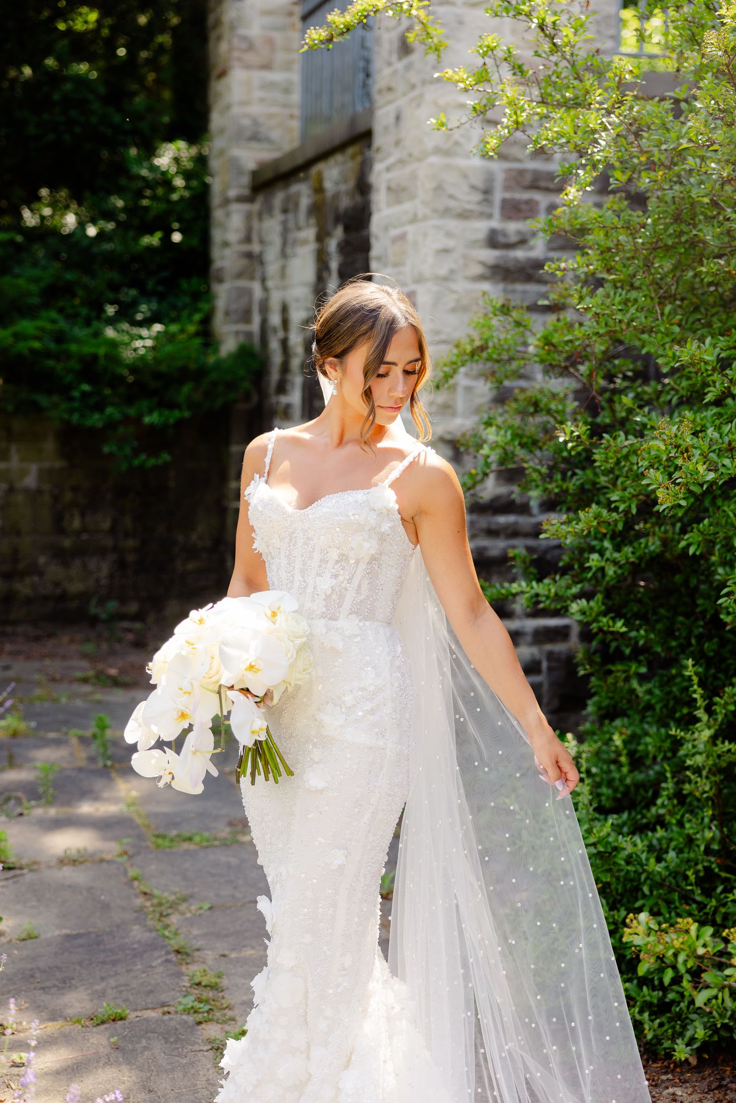 A bride in a white lace wedding dress with delicate details holds a bouquet of white orchids near a stone church wall.