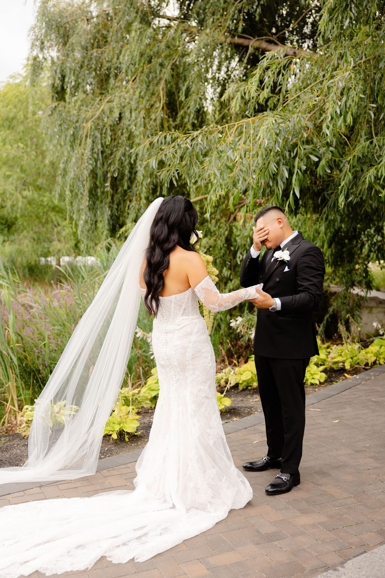 Two people share a candid moment during a wedding ceremony under a willow tree.