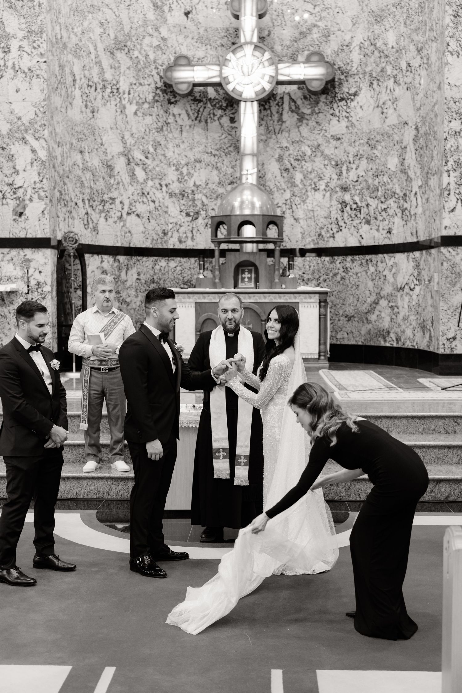 A black and white photo of a wedding ceremony with attendants adjusting the bride's dress.