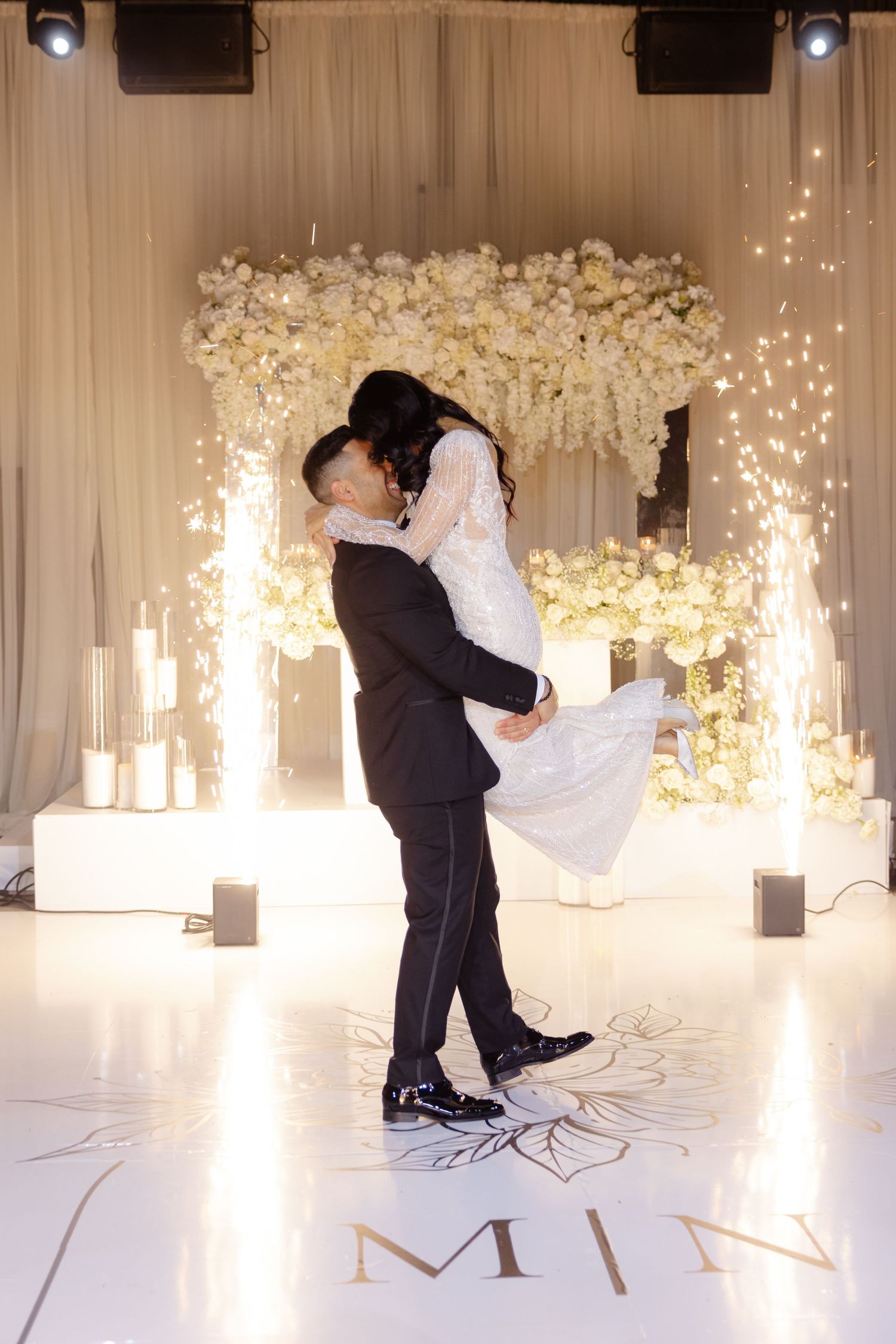 Wedding couple shares intimate dance moment in front of illuminated white flower wall with cold sparklers.