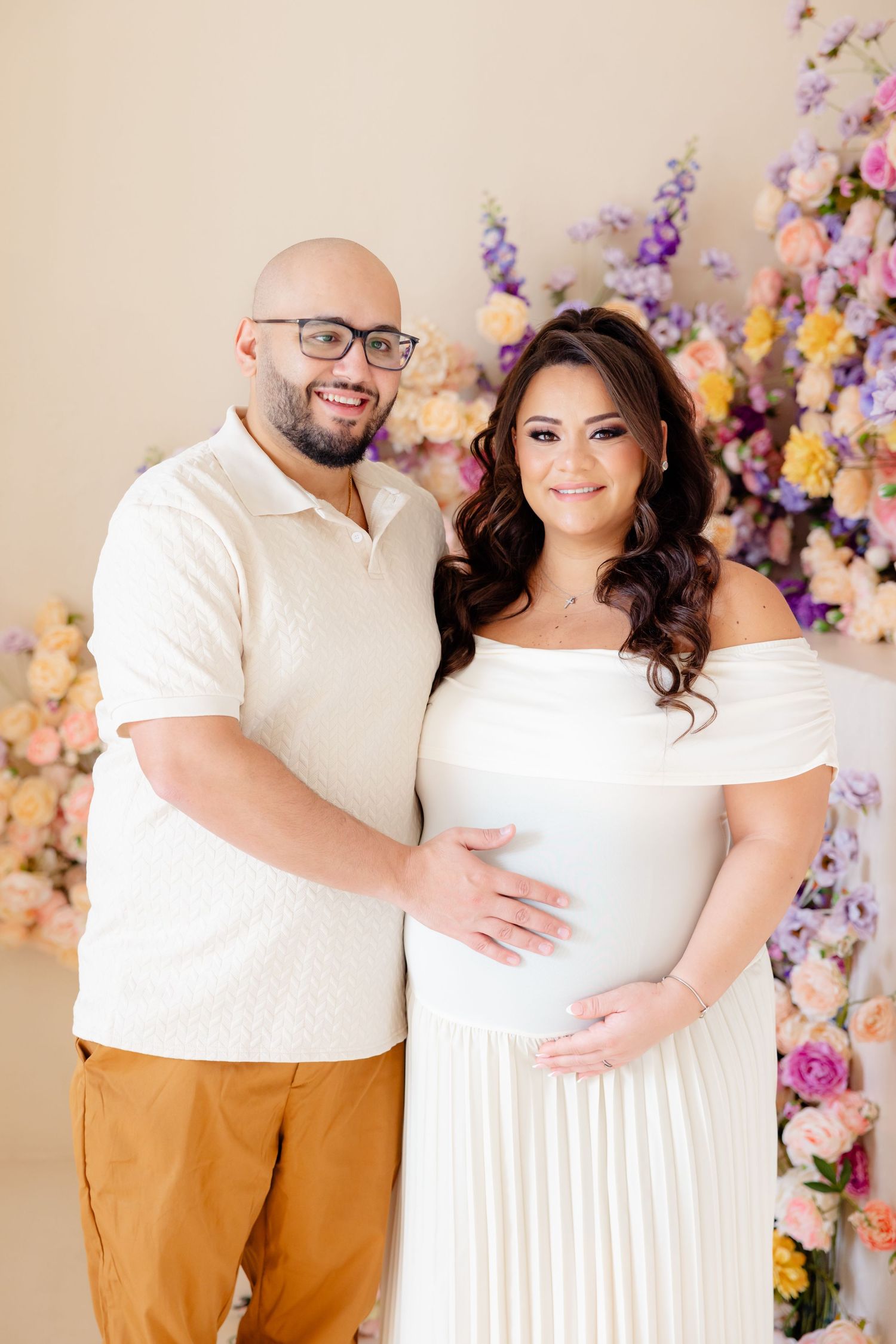 A couple poses against a floral backdrop while celebrating their pregnancy in matching white outfits.