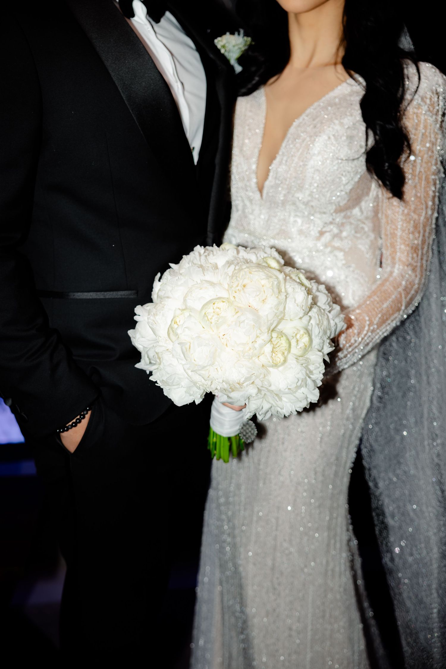 Close-up of a white peony bouquet held by a bride in a sparkly wedding gown next to a groom in a tuxedo.