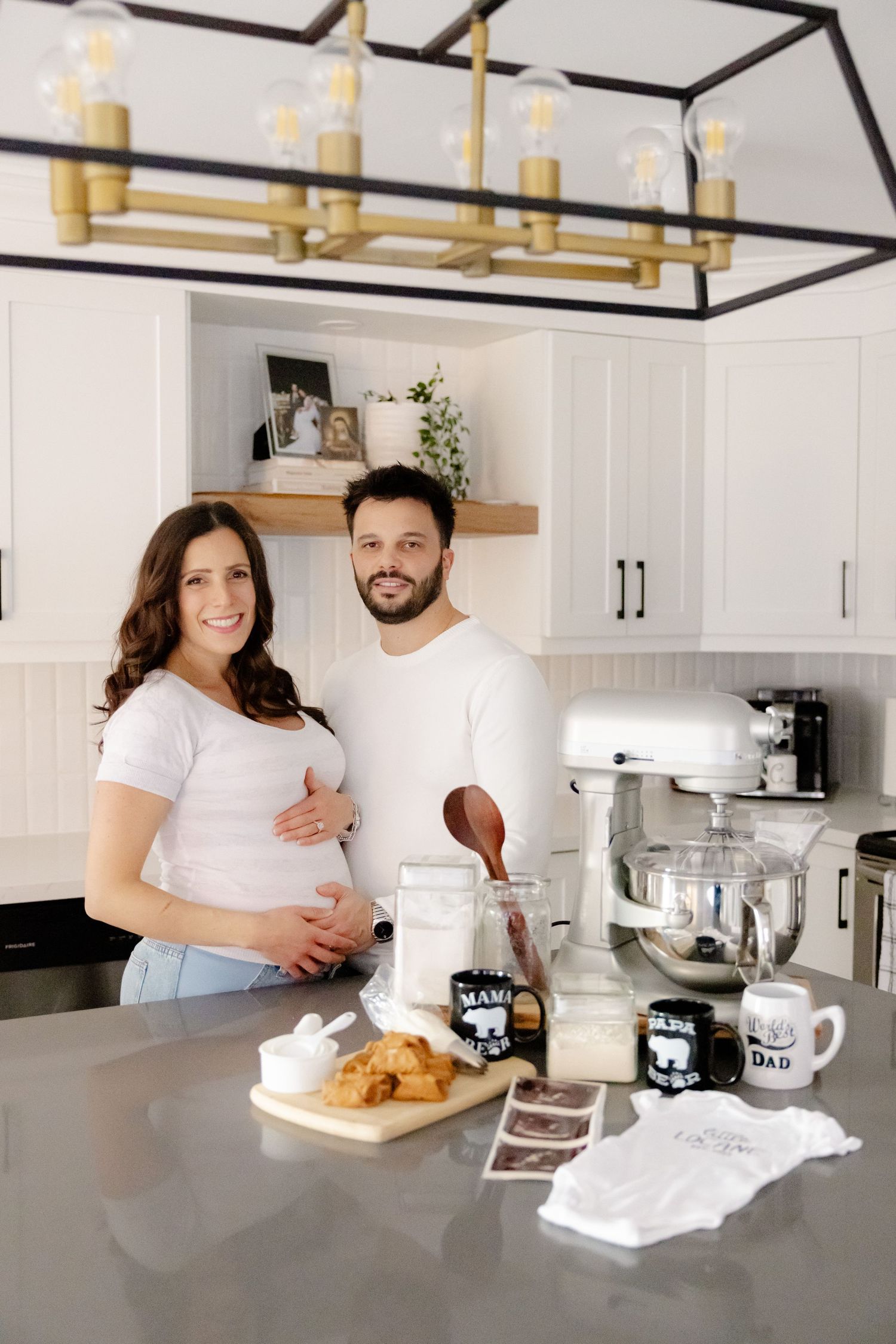 A couple stands at their kitchen counter with baked goods and coffee mugs, under a modern black and brass light fixture.