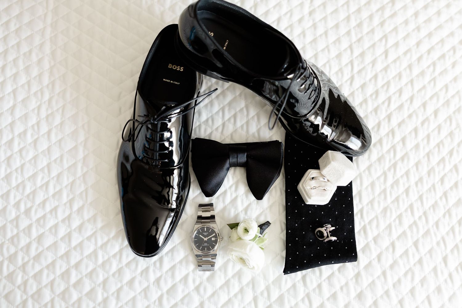 Black patent leather dress shoes arranged with a bow tie, silver watch, and designer accessories on white textured surface.