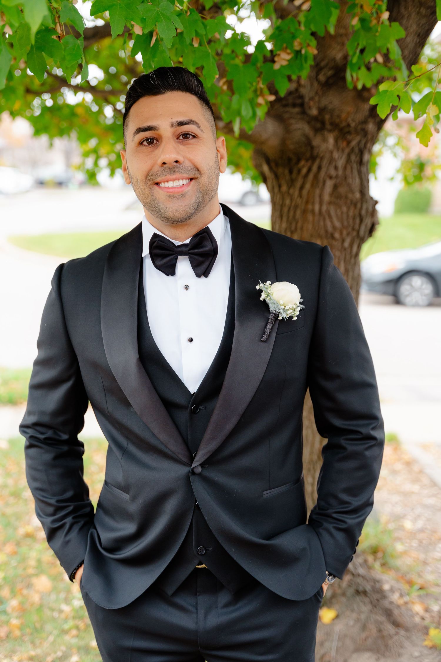 Sharp dressed groom in black tuxedo with bow tie and boutonniere smiling outdoors.