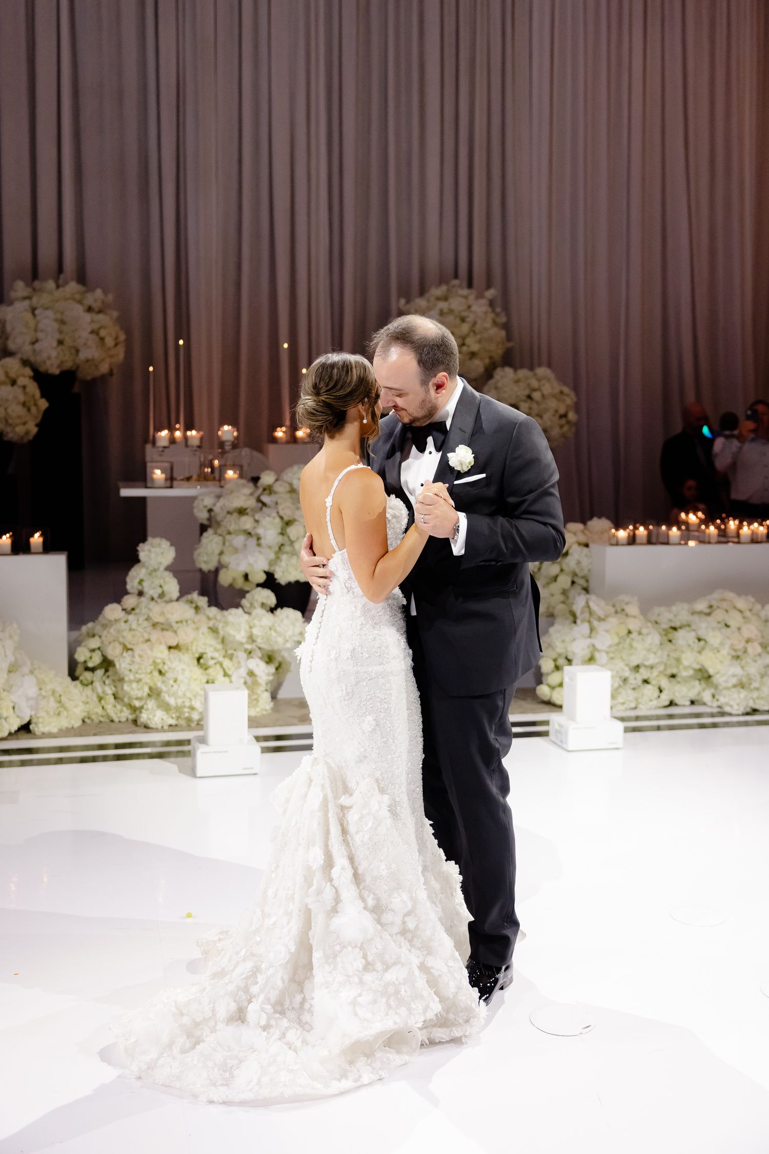 A couple shares their first dance at a wedding reception with a white floral backdrop and soft candlelight.