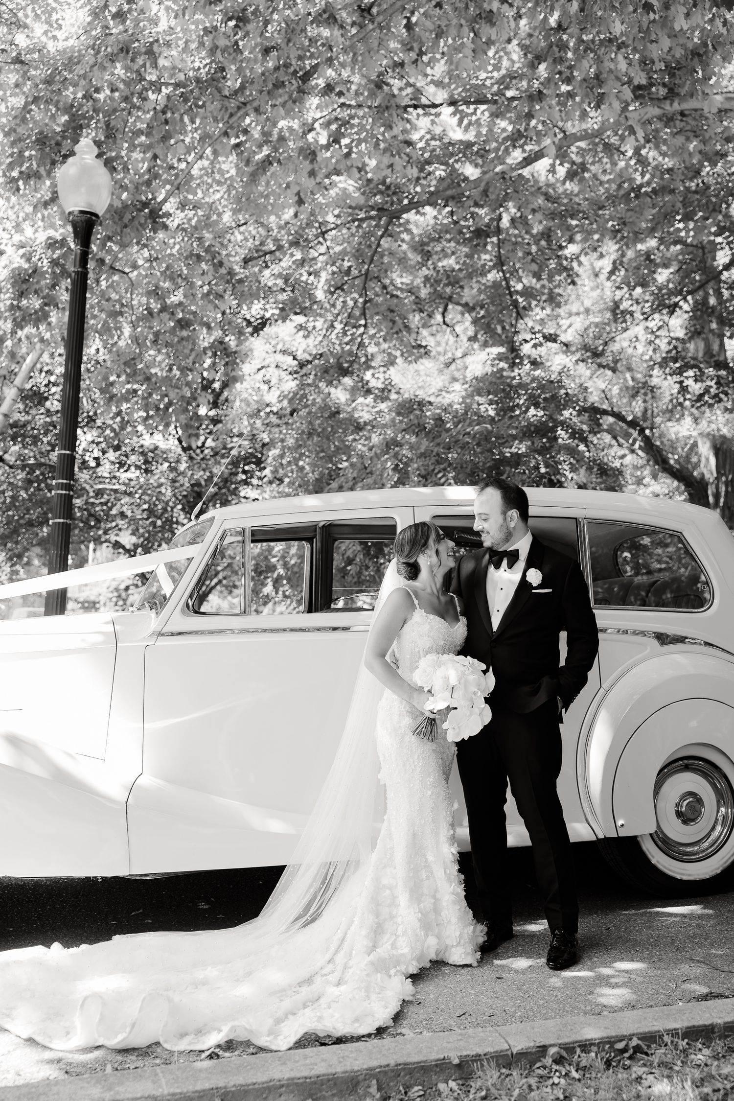 A classic white vintage car serves as an elegant backdrop for a romantic wedding photo under tree canopy.