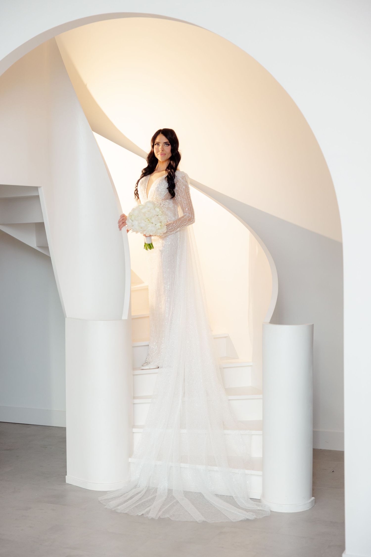 Elegant bridal portrait on curved white staircase holding white bouquet.