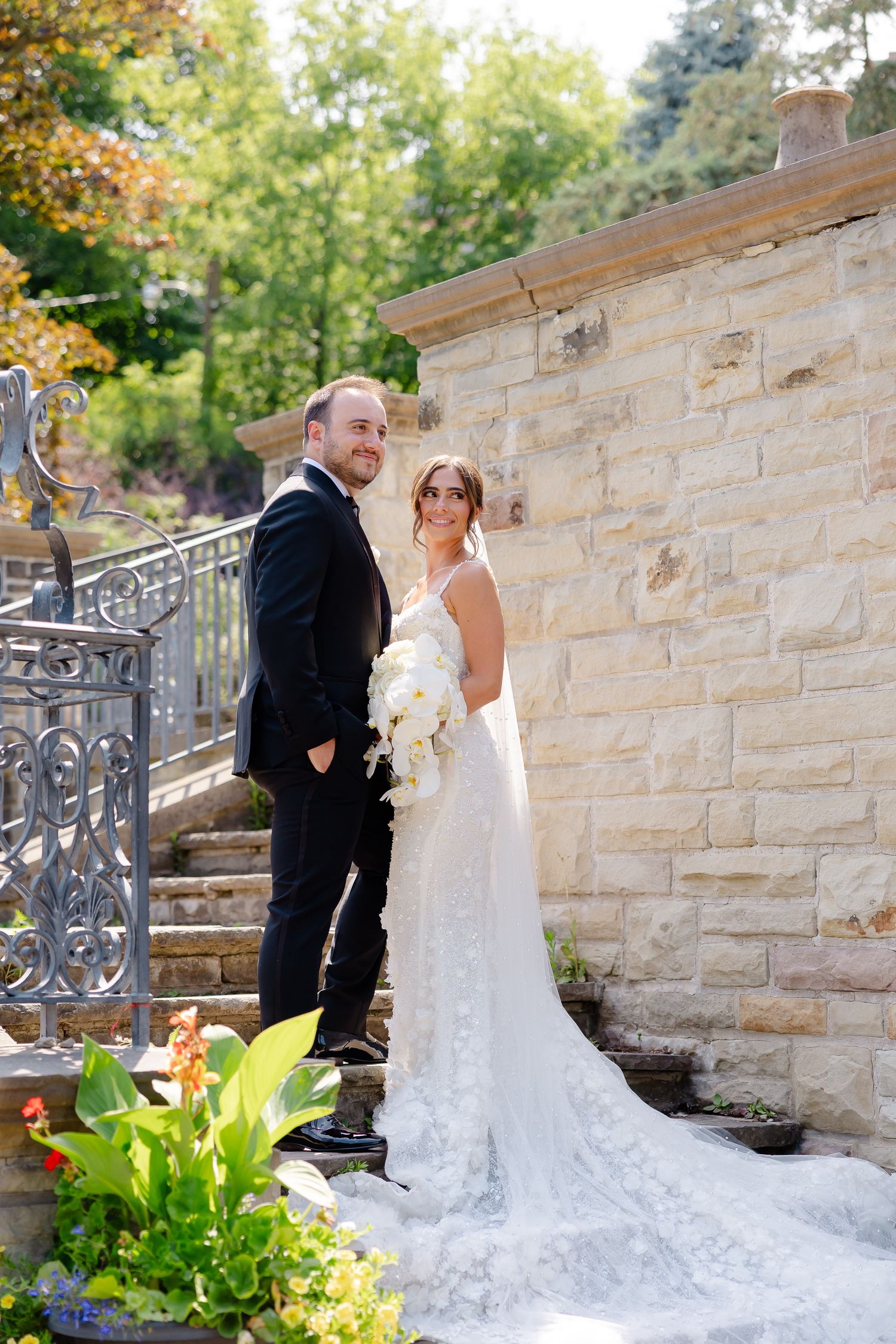 A couple poses on stone steps next to an ornate railing and flowering plants on their wedding day.