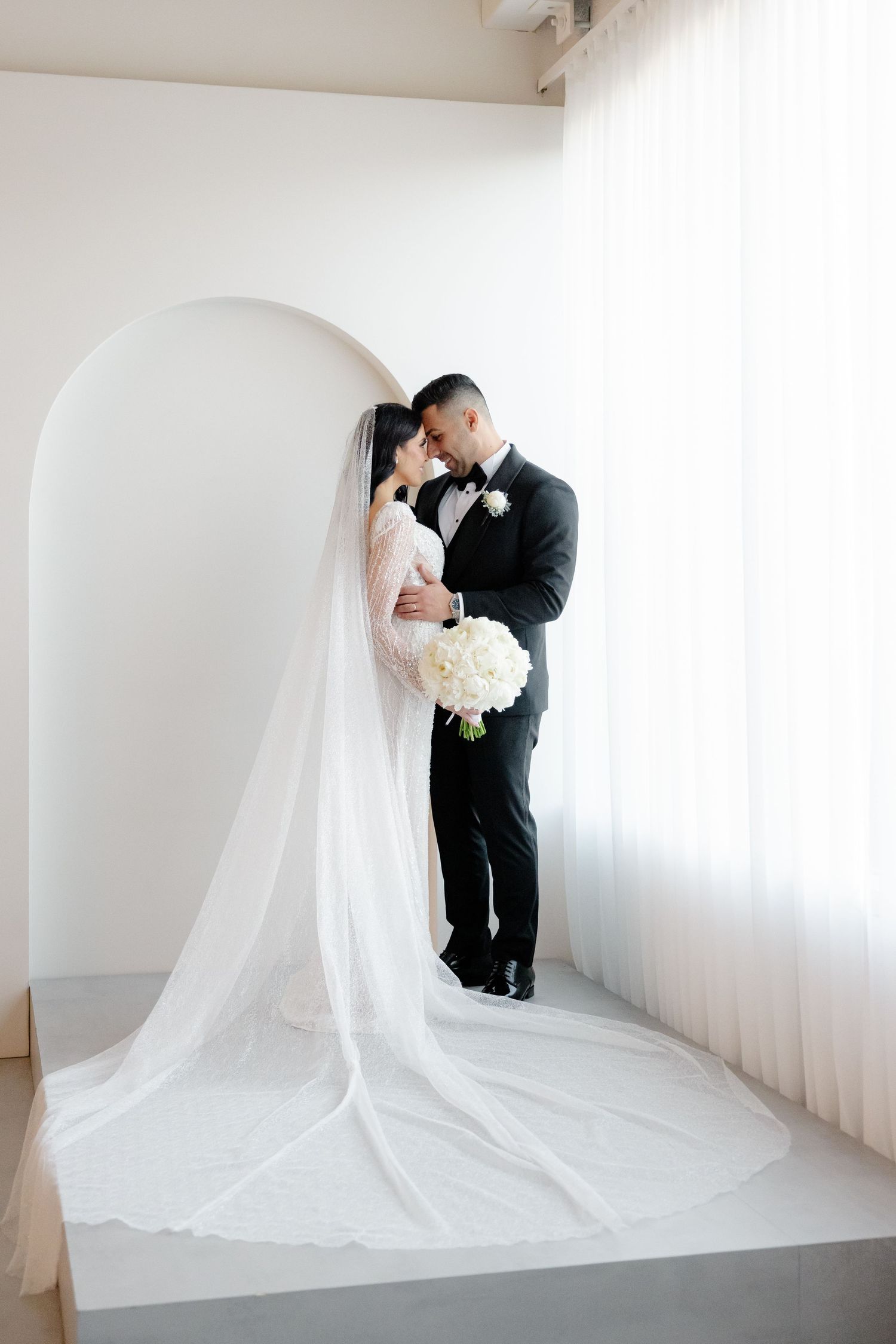 A romantic wedding portrait with couple embracing under white archway with dramatic veil.