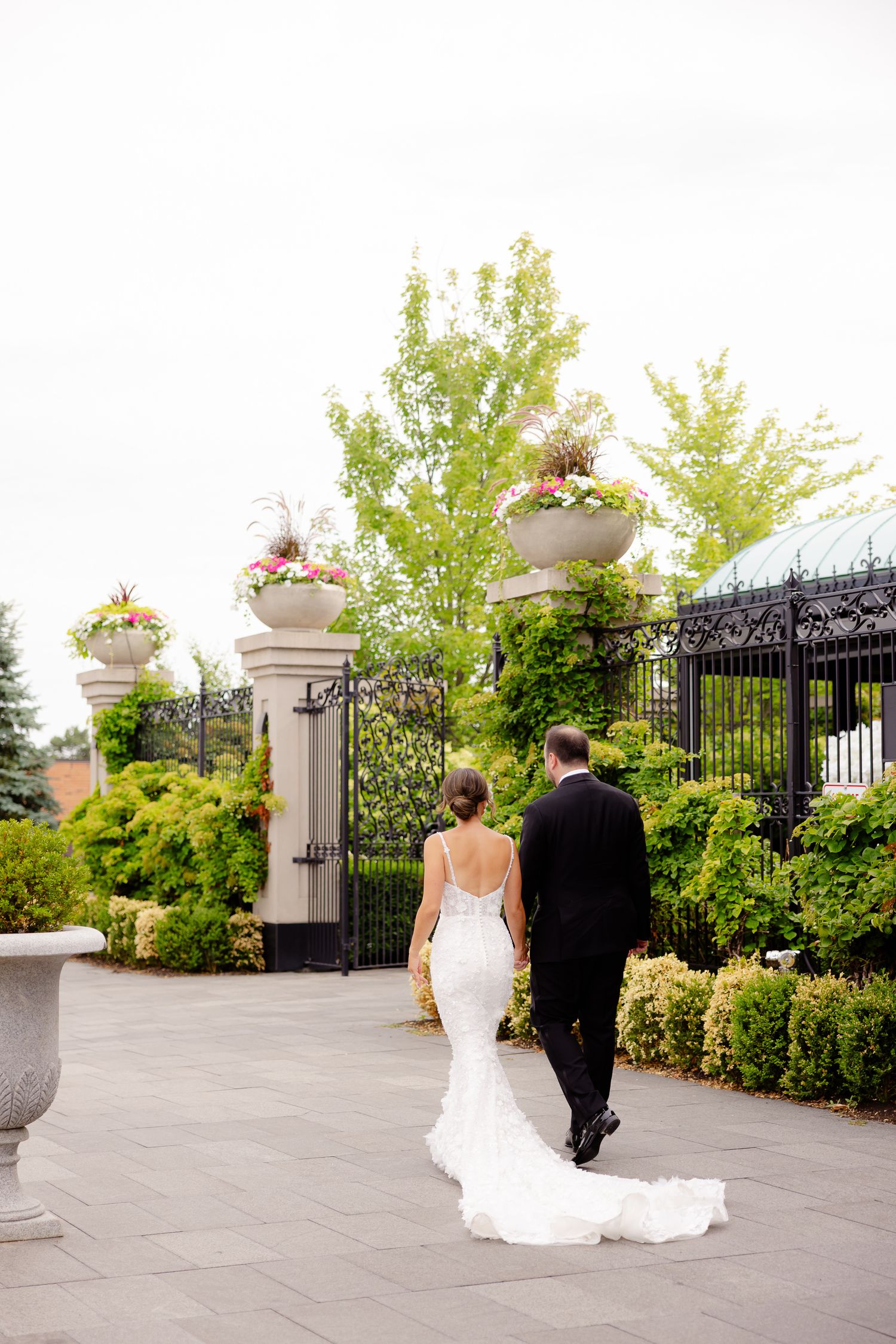 A couple walks through an elegant garden terrace with decorative stone pillars and flowering urns at an outdoor venue.