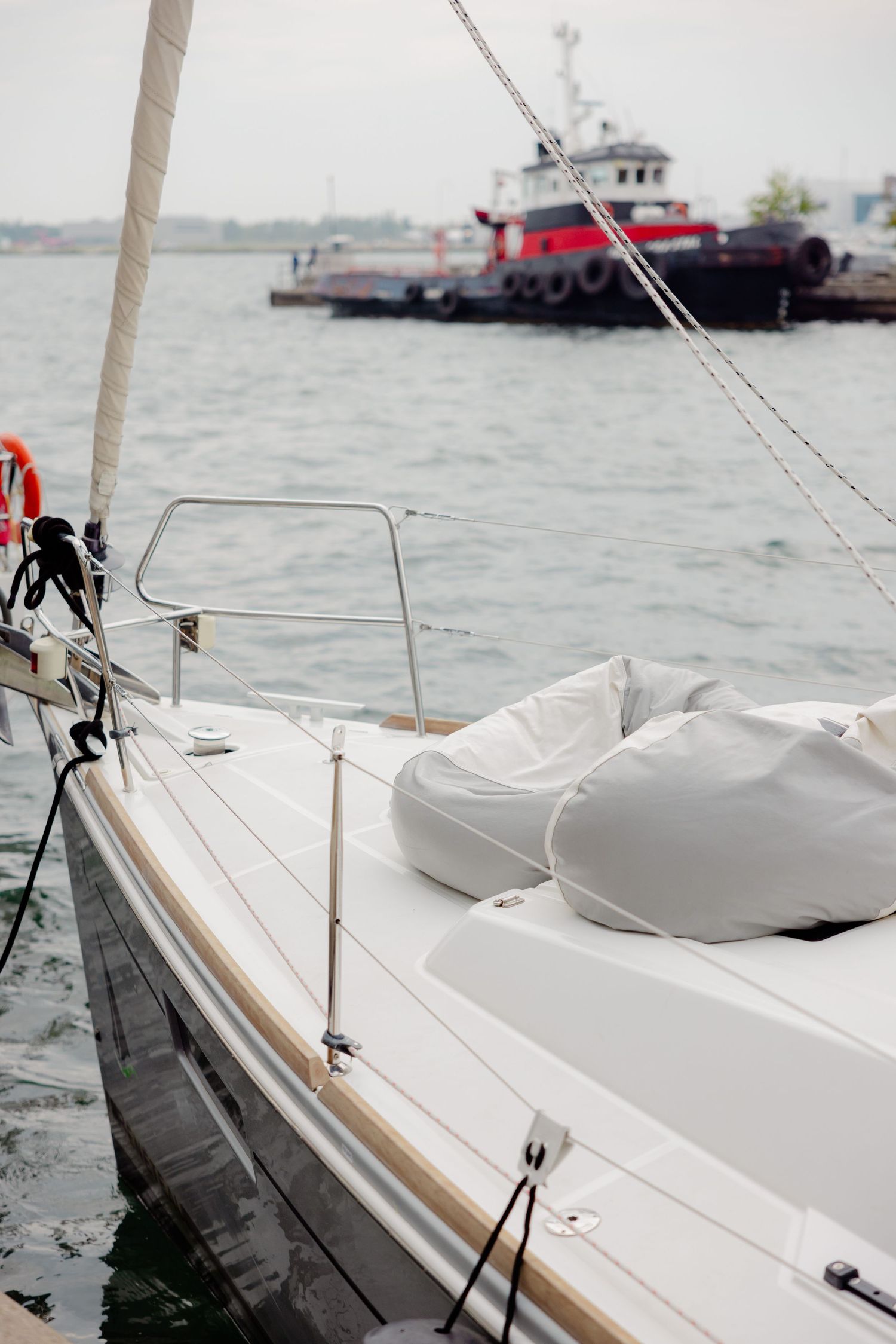 Side view of a white sailboat deck with ropes and rigging, with a red tugboat visible in the background on choppy waters.