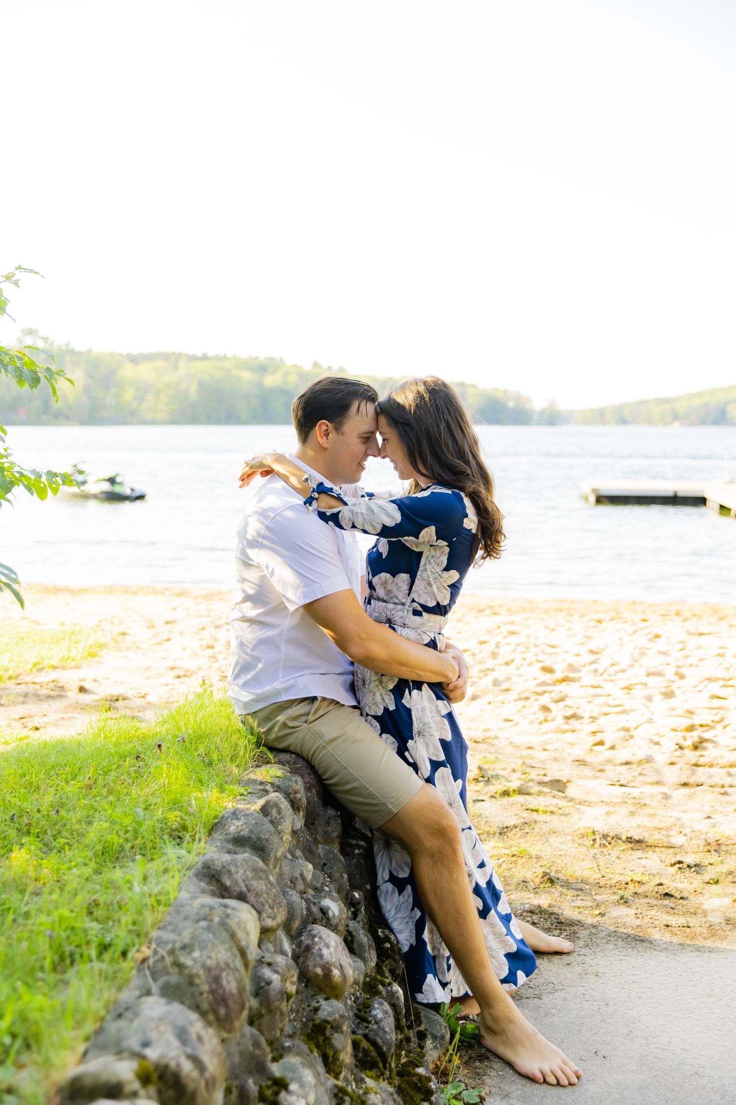 A romantic waterfront portrait of a couple embracing on a stone wall with a lake in the background.