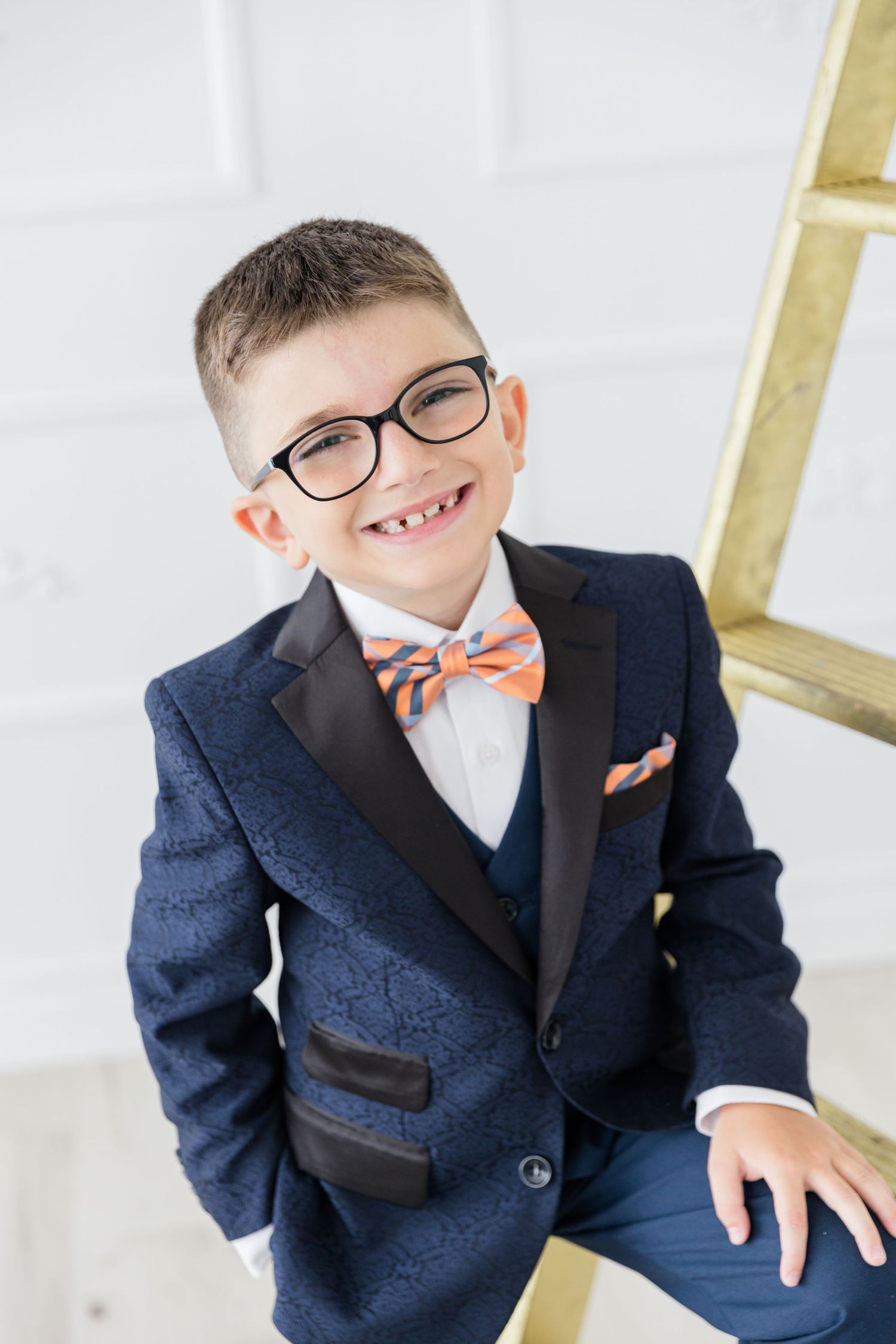 Portrait shot of youth in formal navy suit with orange bowtie near gold ladder.
