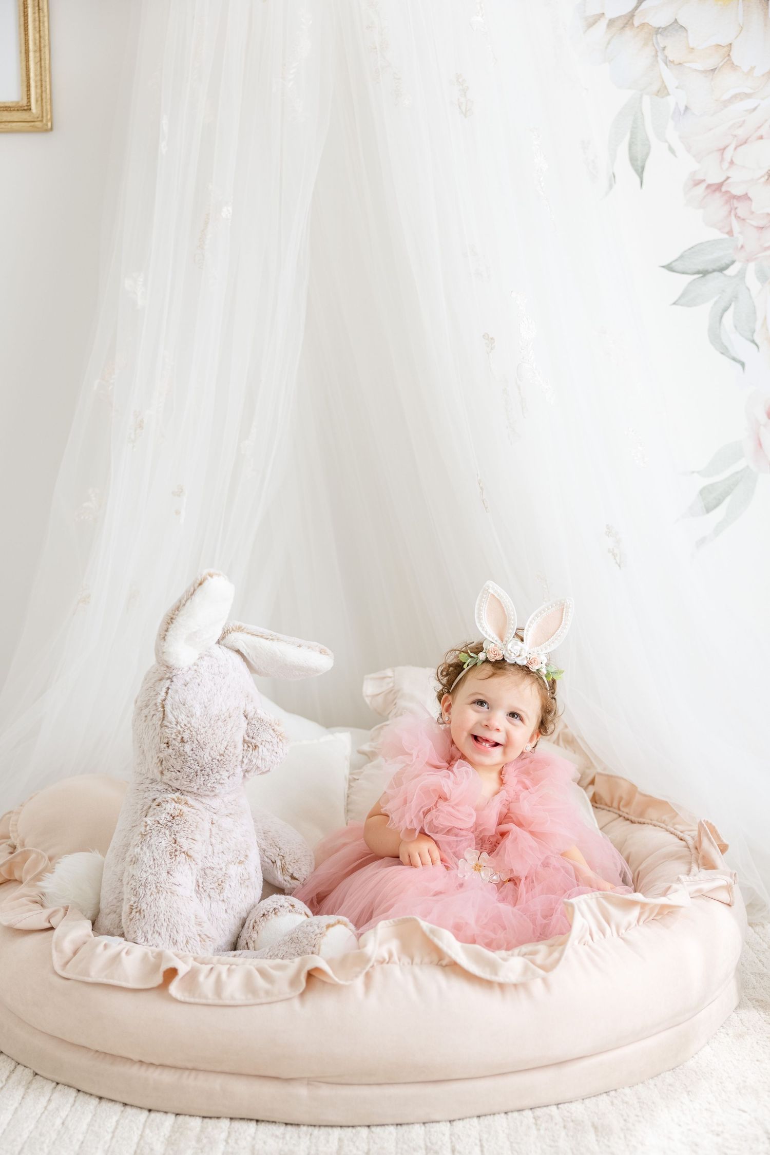 A toddler wearing pink dress and bunny ears sits beside white bunny decor on round cushioned platform.