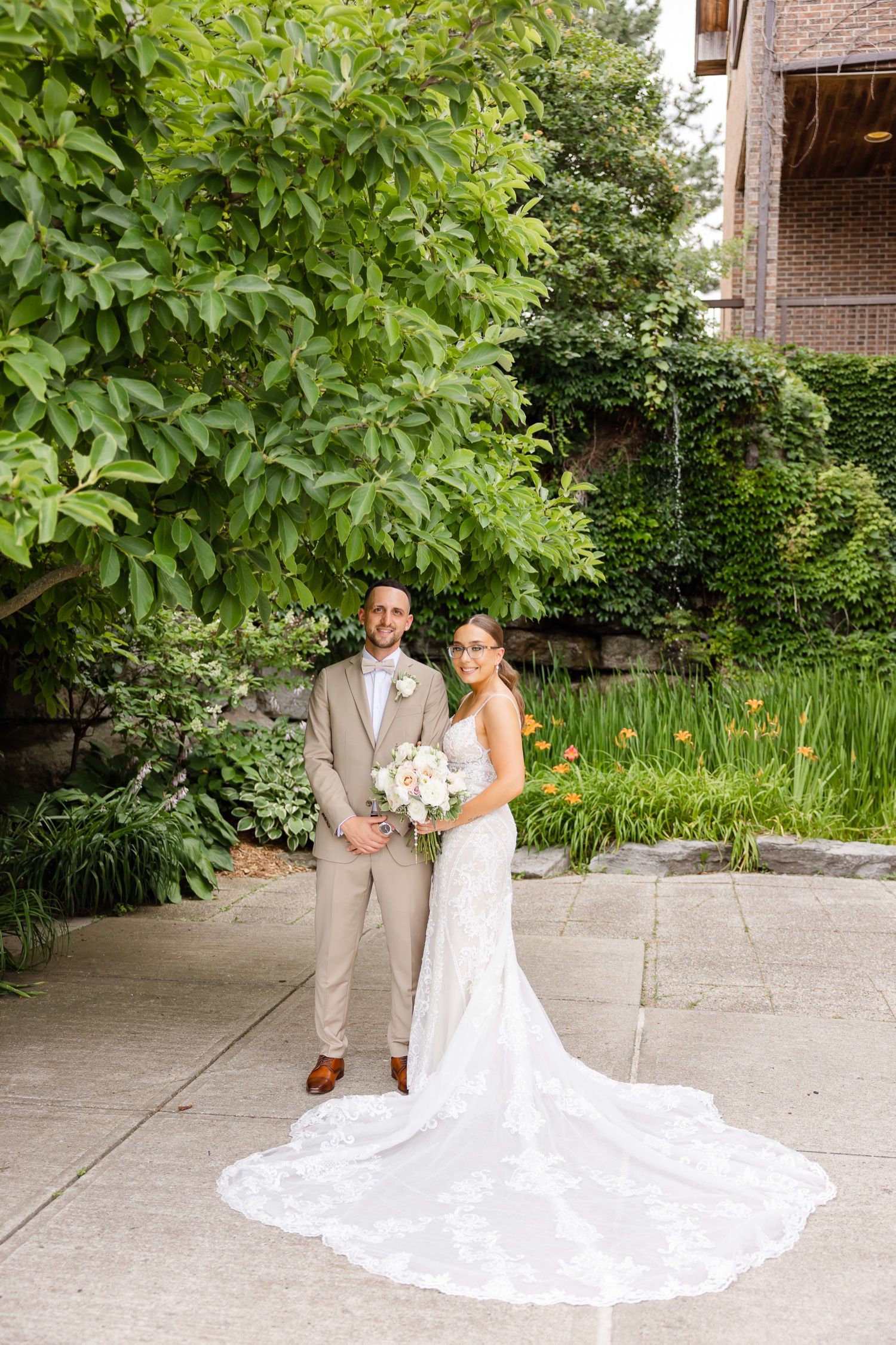 Elegant wedding photo featuring long lace train against garden landscape and brick building.
