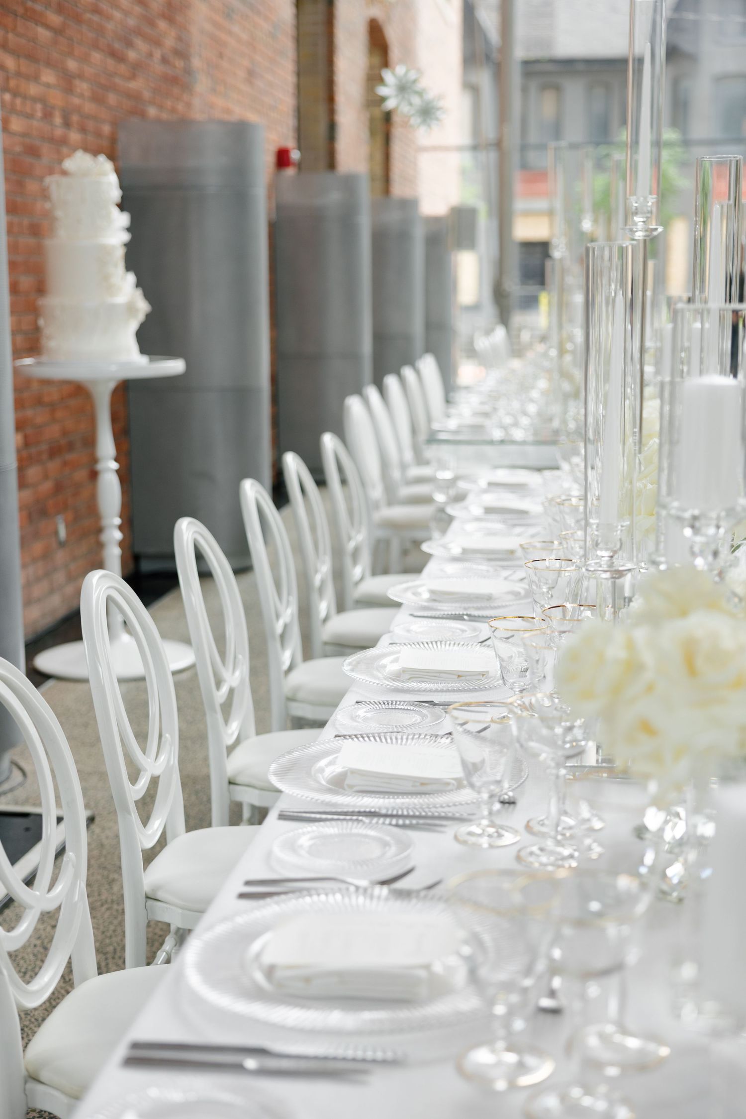 White dining chairs line a formal table setting against exposed brick walls with tall candle holders.