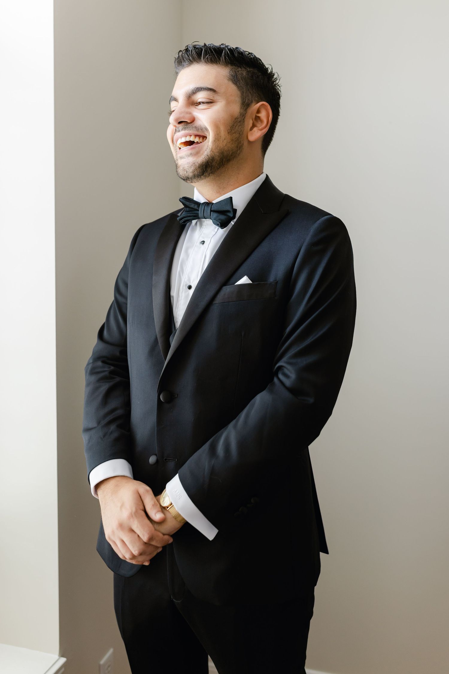 Groom in classic black tuxedo and bow tie against white wall.