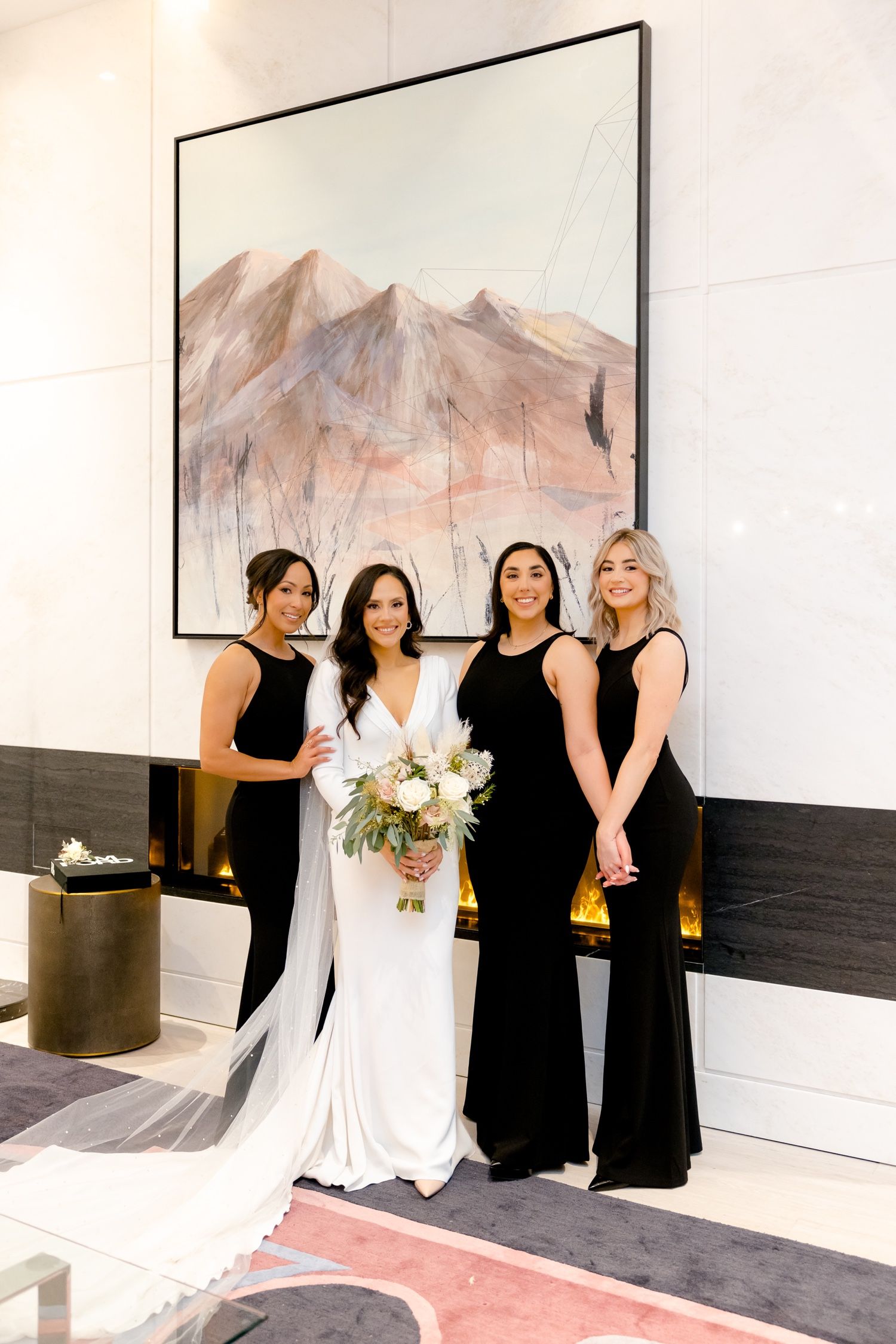 Bridal party poses elegantly in black gowns with bride in white against mountain artwork.