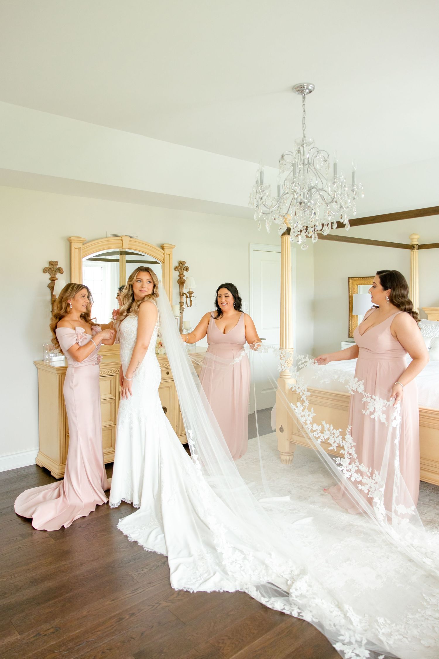 Bridal party in blush pink dresses admiring wedding gown details under crystal chandelier.