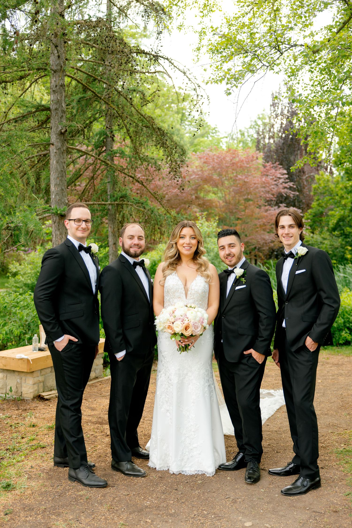 Wedding party in formal attire poses together on garden path with trees in background.