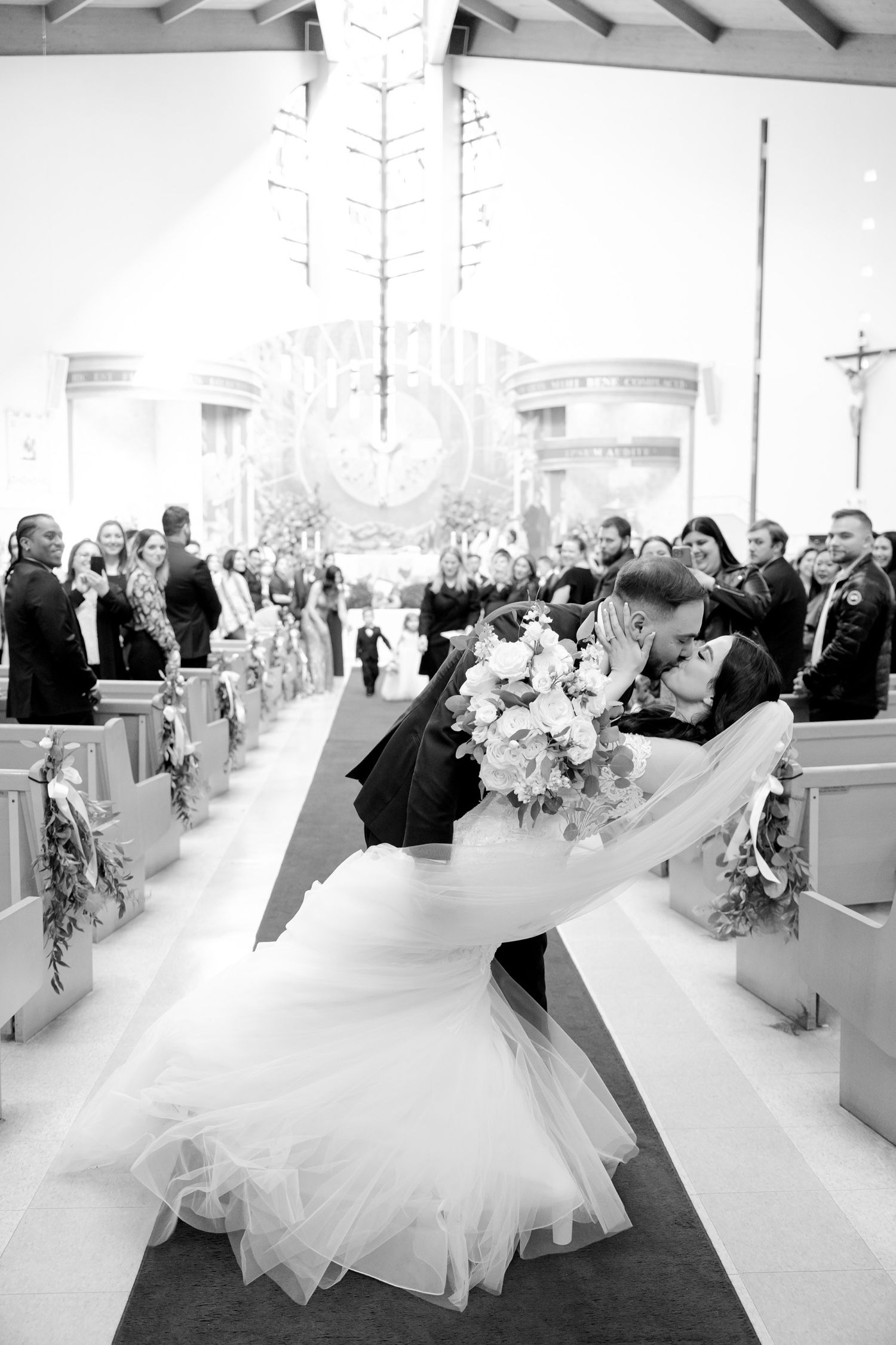 Dramatic black and white photo of newlyweds sharing romantic dip kiss in church aisle.