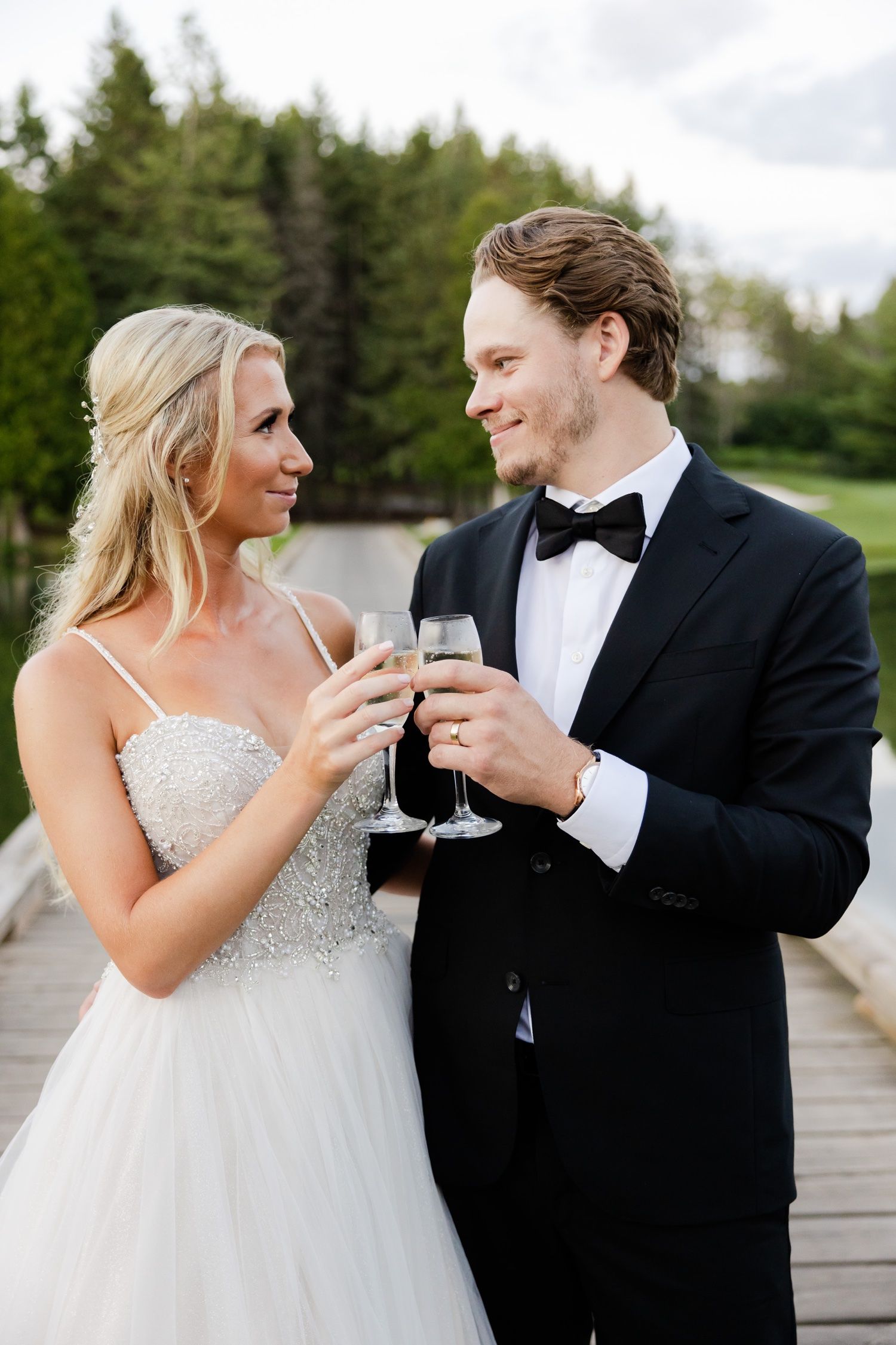 A couple shares champagne and smiles during their wedding celebration on a lakeside dock.