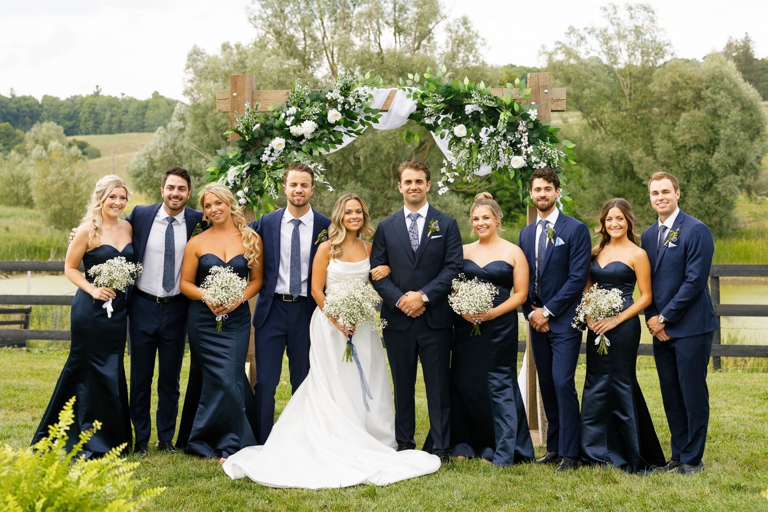 Formal group photo of wedding party in navy and blue attire under floral arch outdoors.
