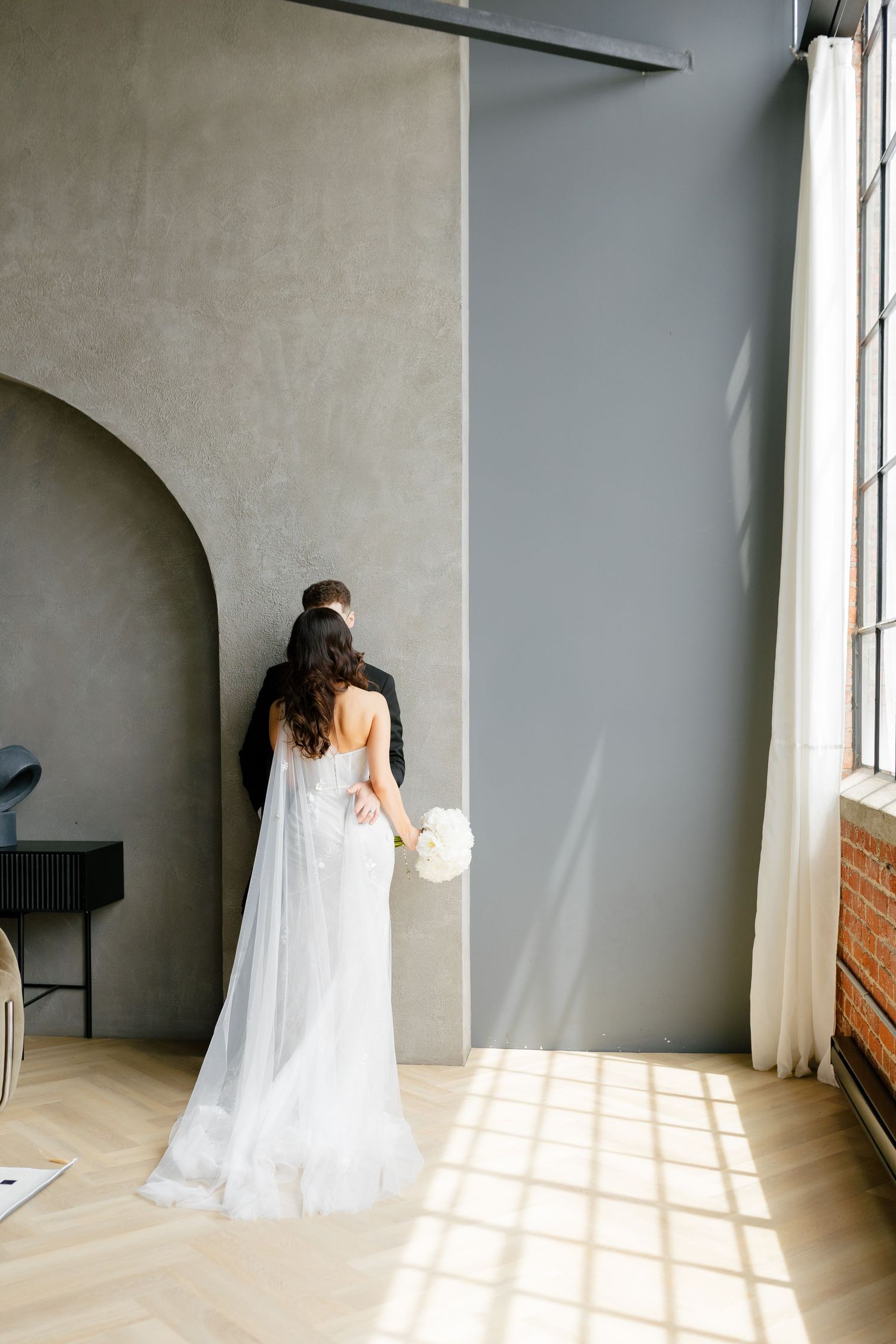 Bride in flowing dress holds bouquet while standing in sunlit room.
