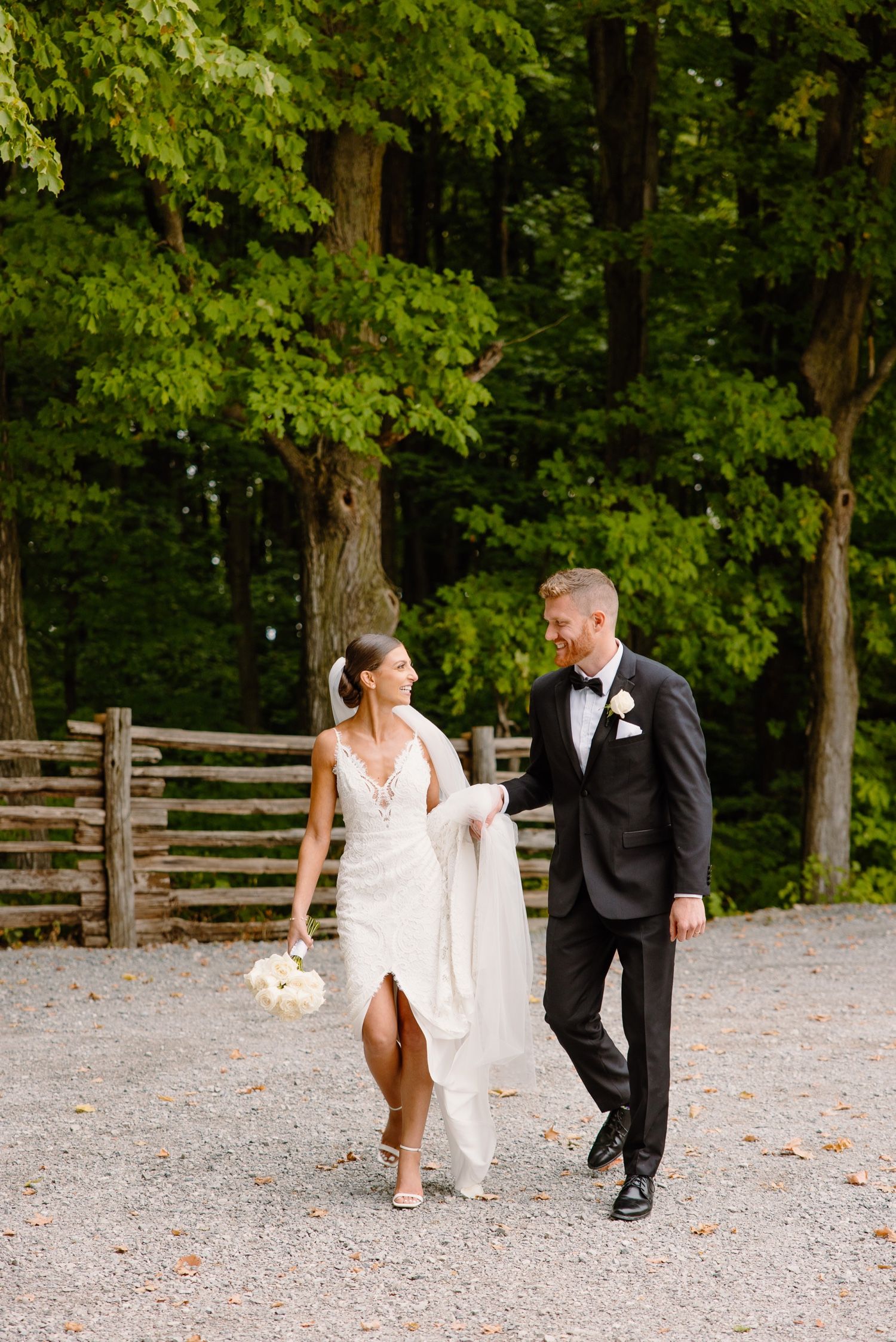 A wedding couple walks along a gravel path with rustic wooden fencing and lush green trees in the background.