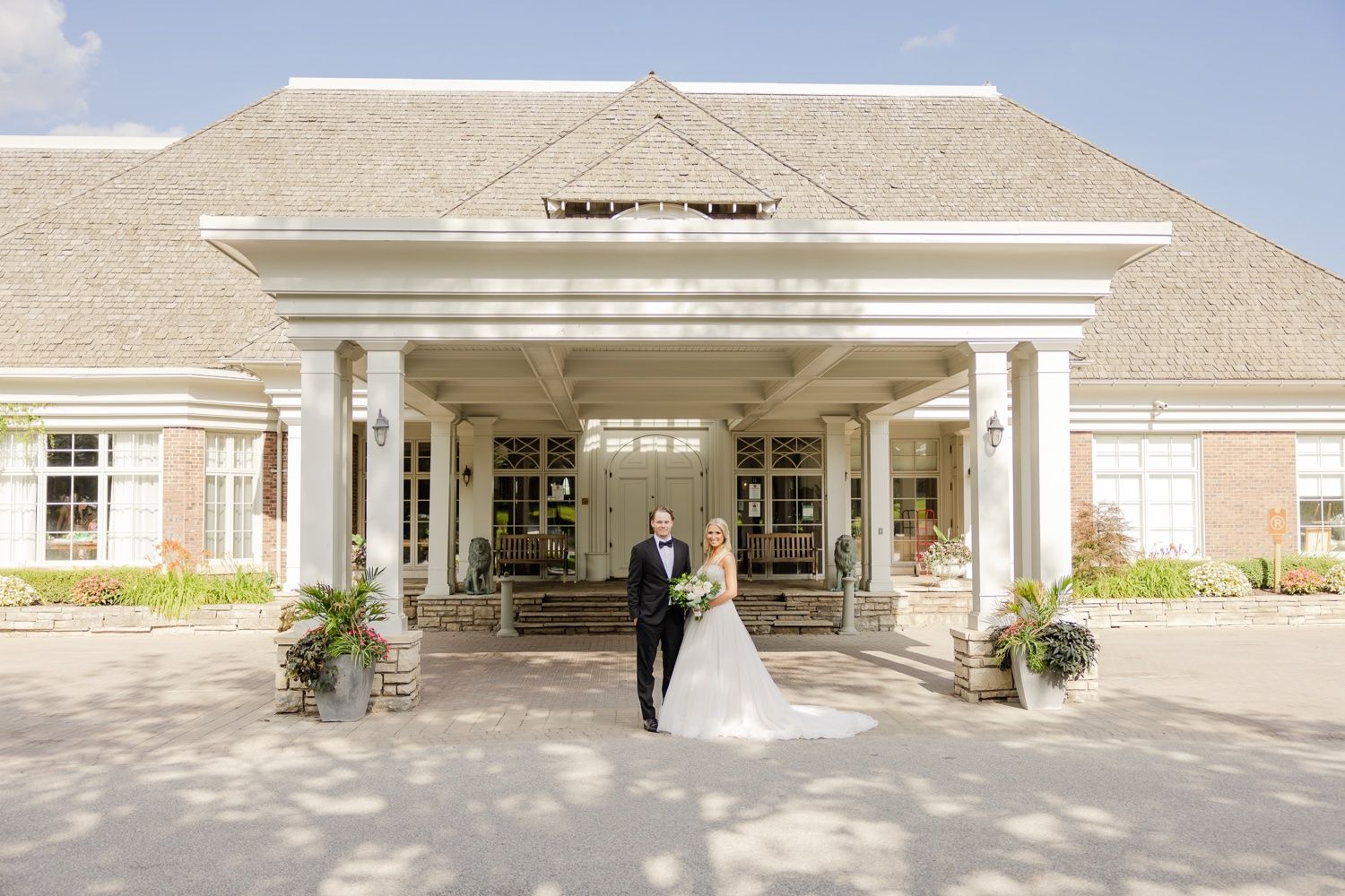 Elegant wedding couple standing under grand white columned entrance of a country club.