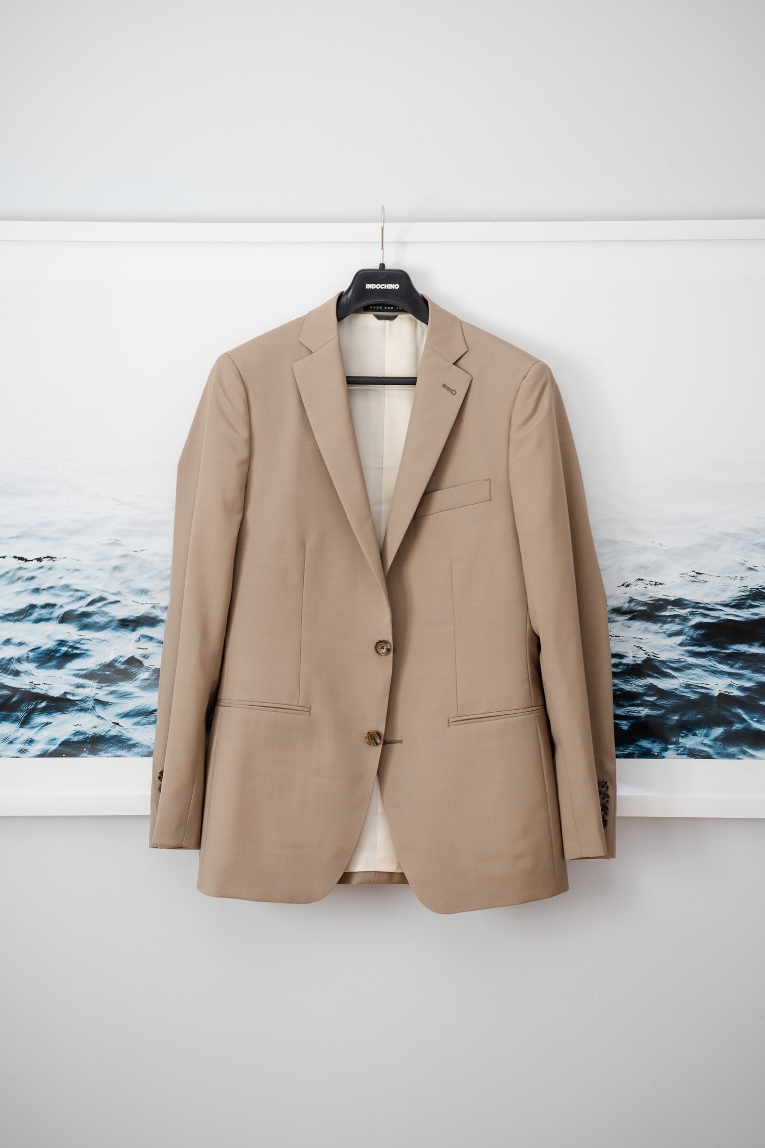 Tan suit jacket displayed on hanger against wall with coastal artwork.