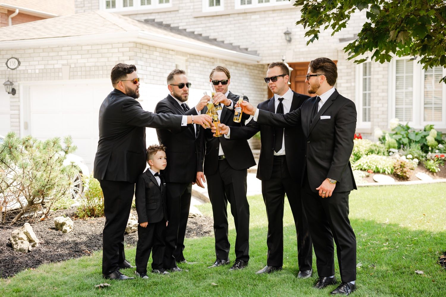Groomsmen in black suits toasting with bottles in outdoor garden setting.