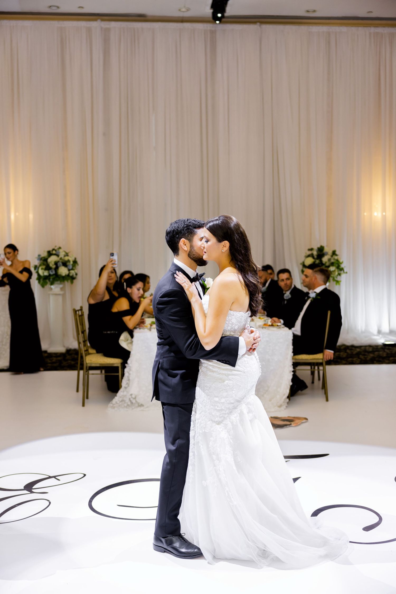 A couple embraces during their first dance on a monogrammed dance floor with white draping and floral decor.