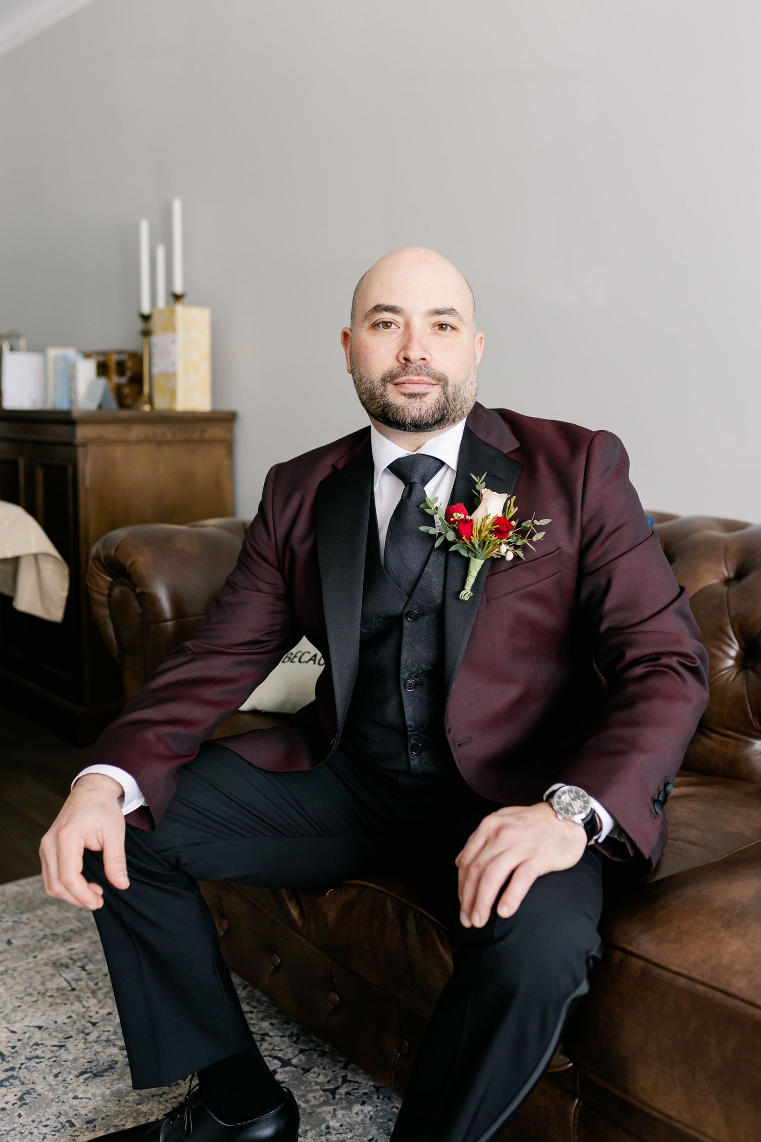 Portrait of groom in burgundy suit sitting on brown leather couch with red boutonniere.