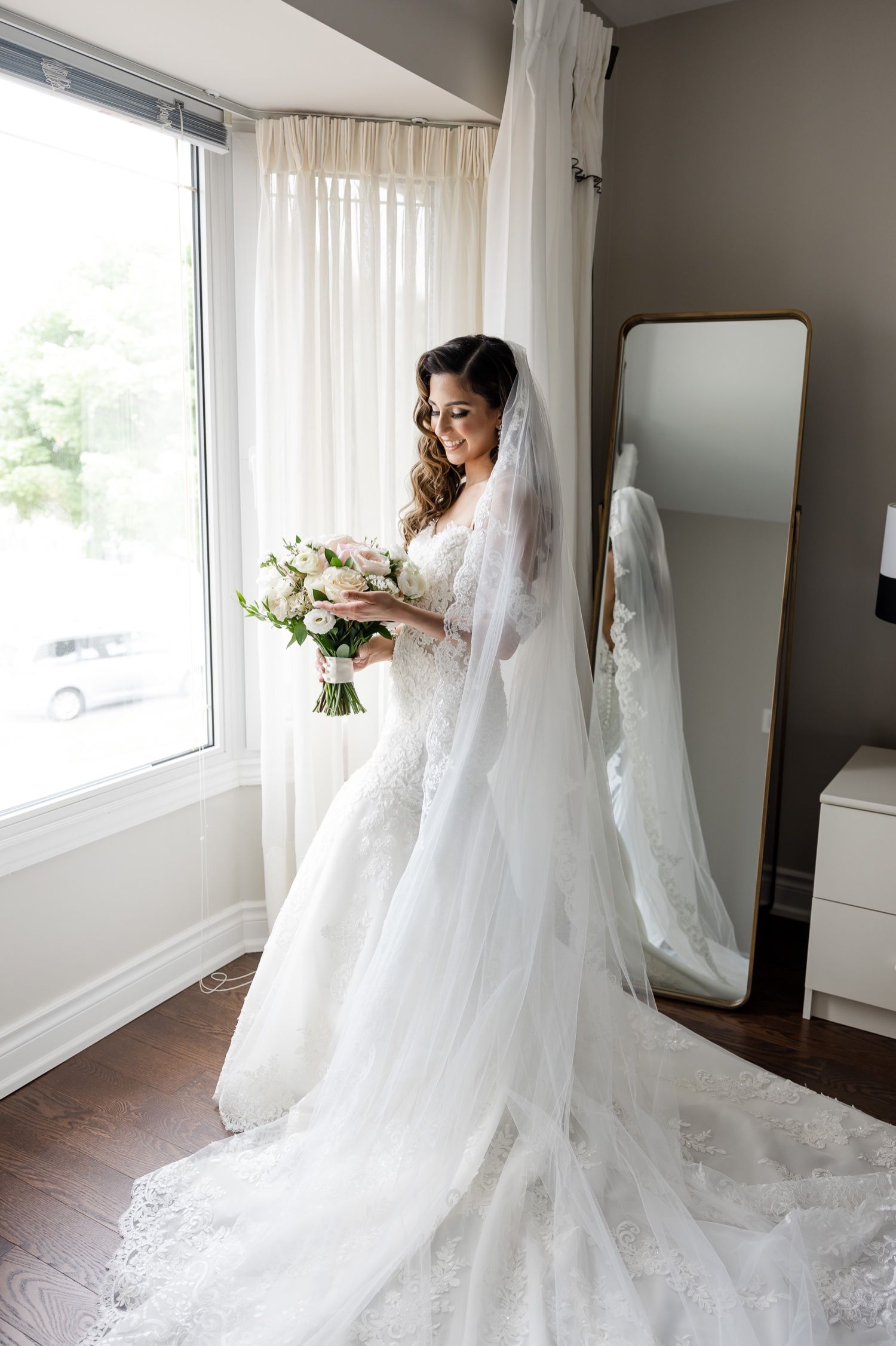 Elegant bride in a lace wedding dress with long veil poses by window holding a pastel bouquet.