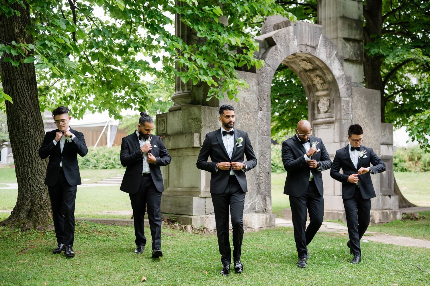 Groomsmen walking in synchronized style wearing black tuxedos through stone archway.