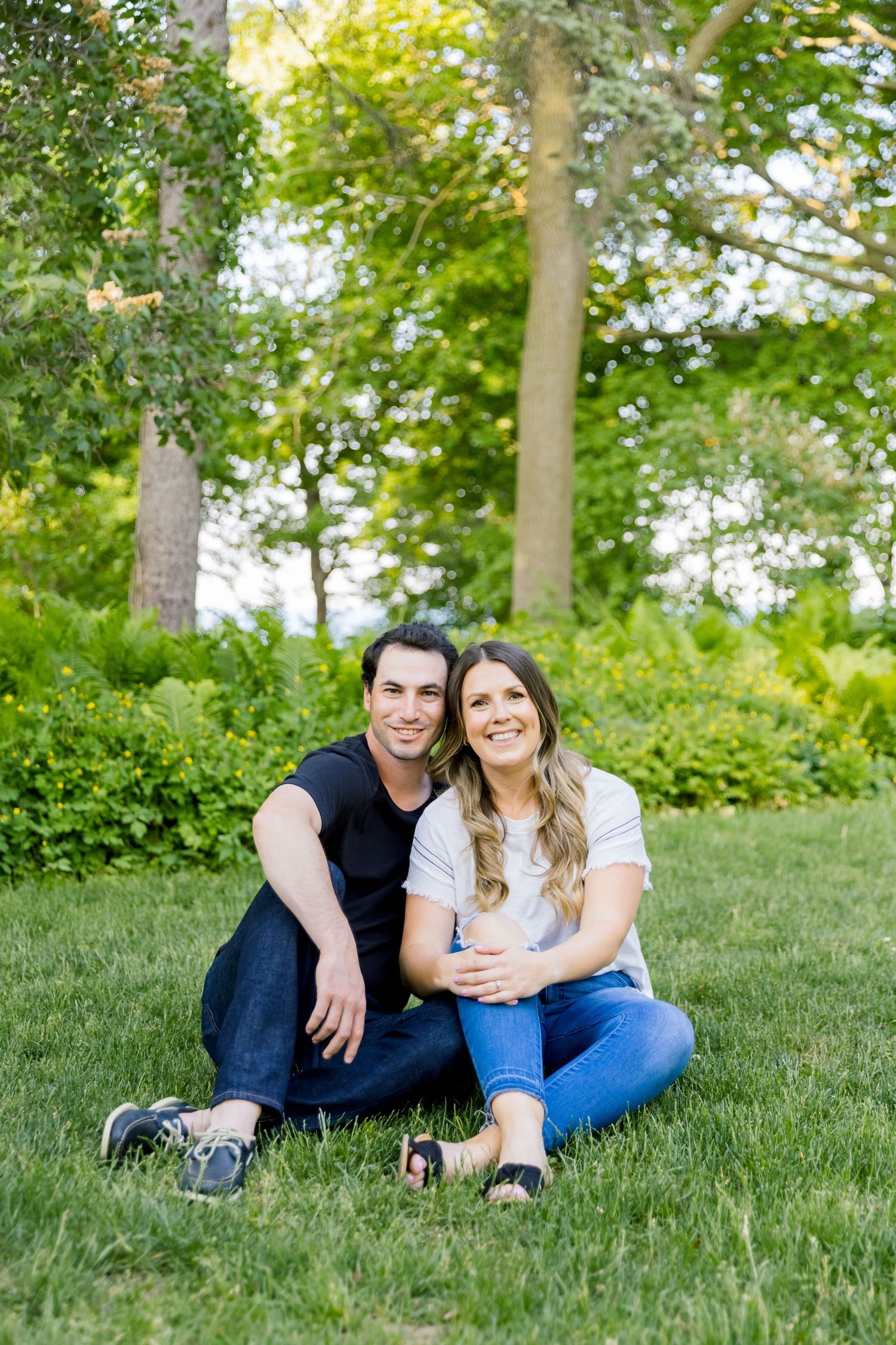 A couple sits together on grass with lush green trees in the background.