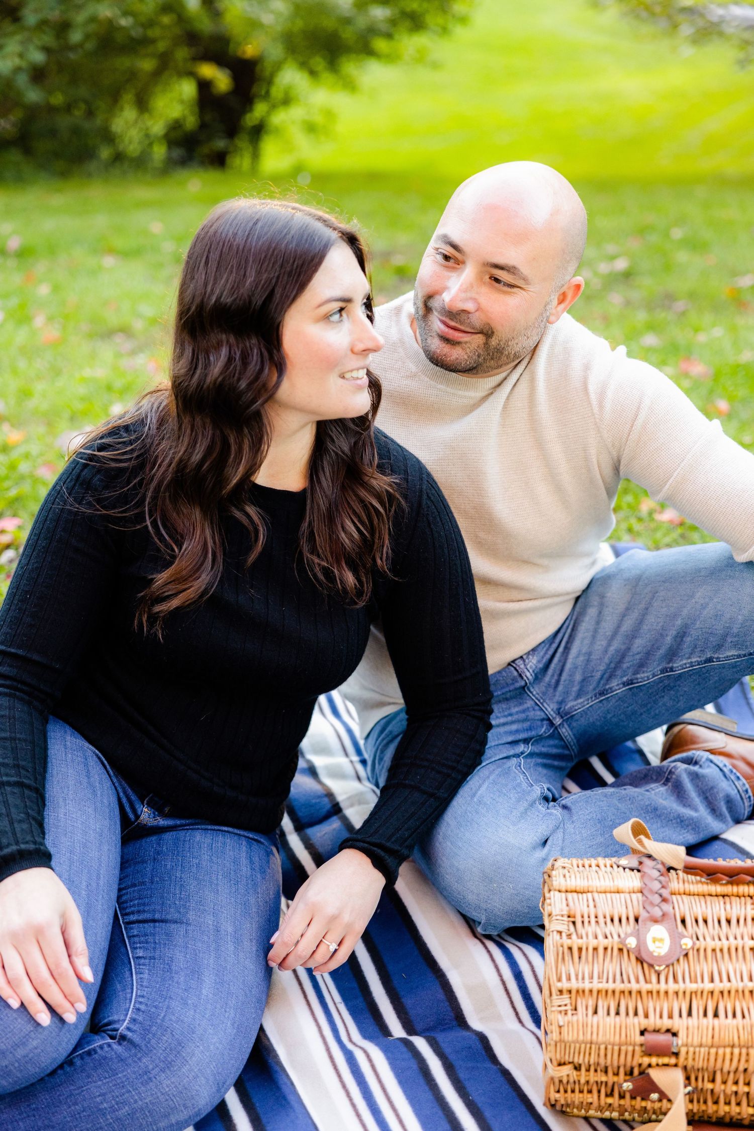 Casual couple sitting close together on picnic blanket with wicker basket nearby.
