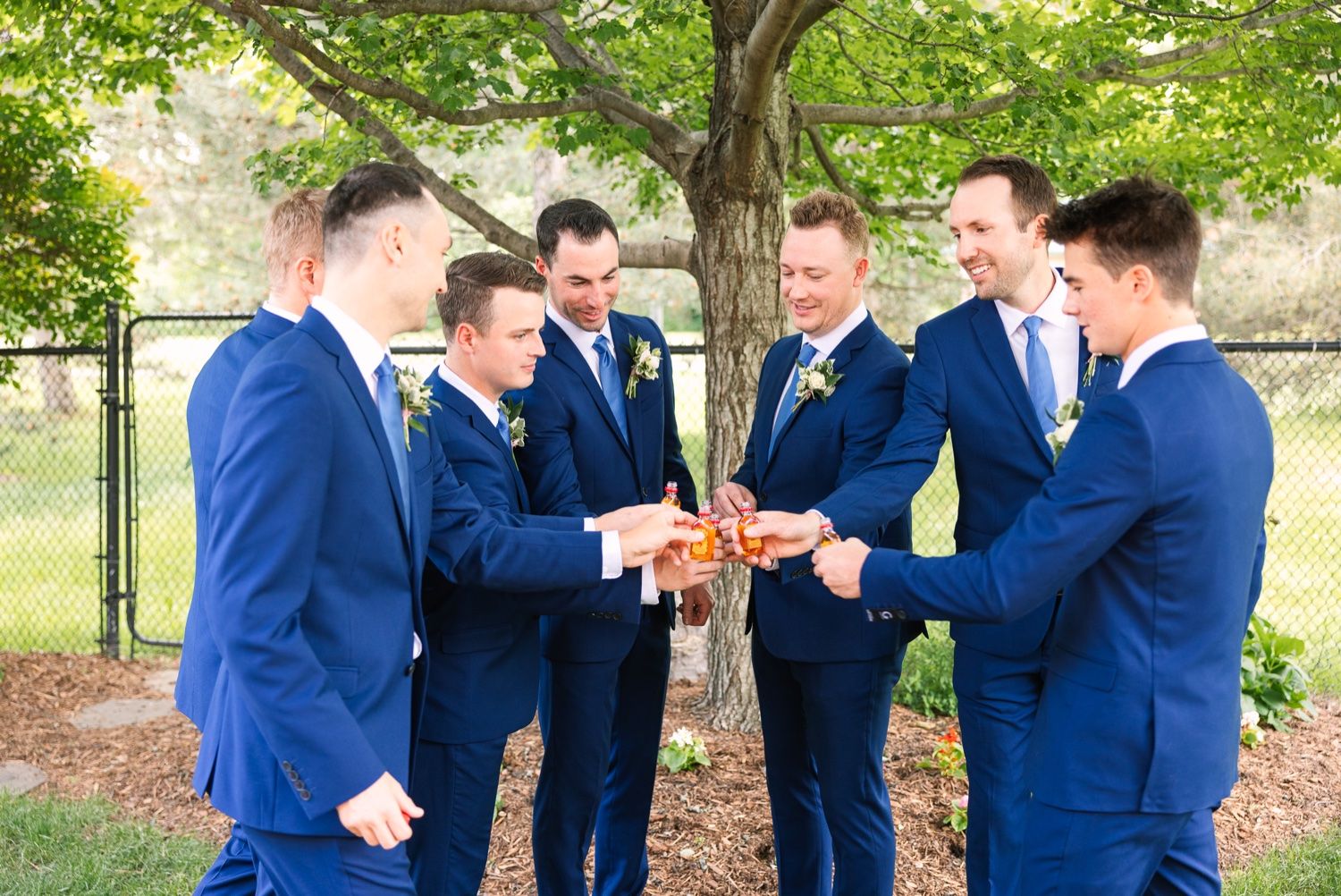 Groomsmen in matching navy blue suits join hands in a circle outdoors under green trees.