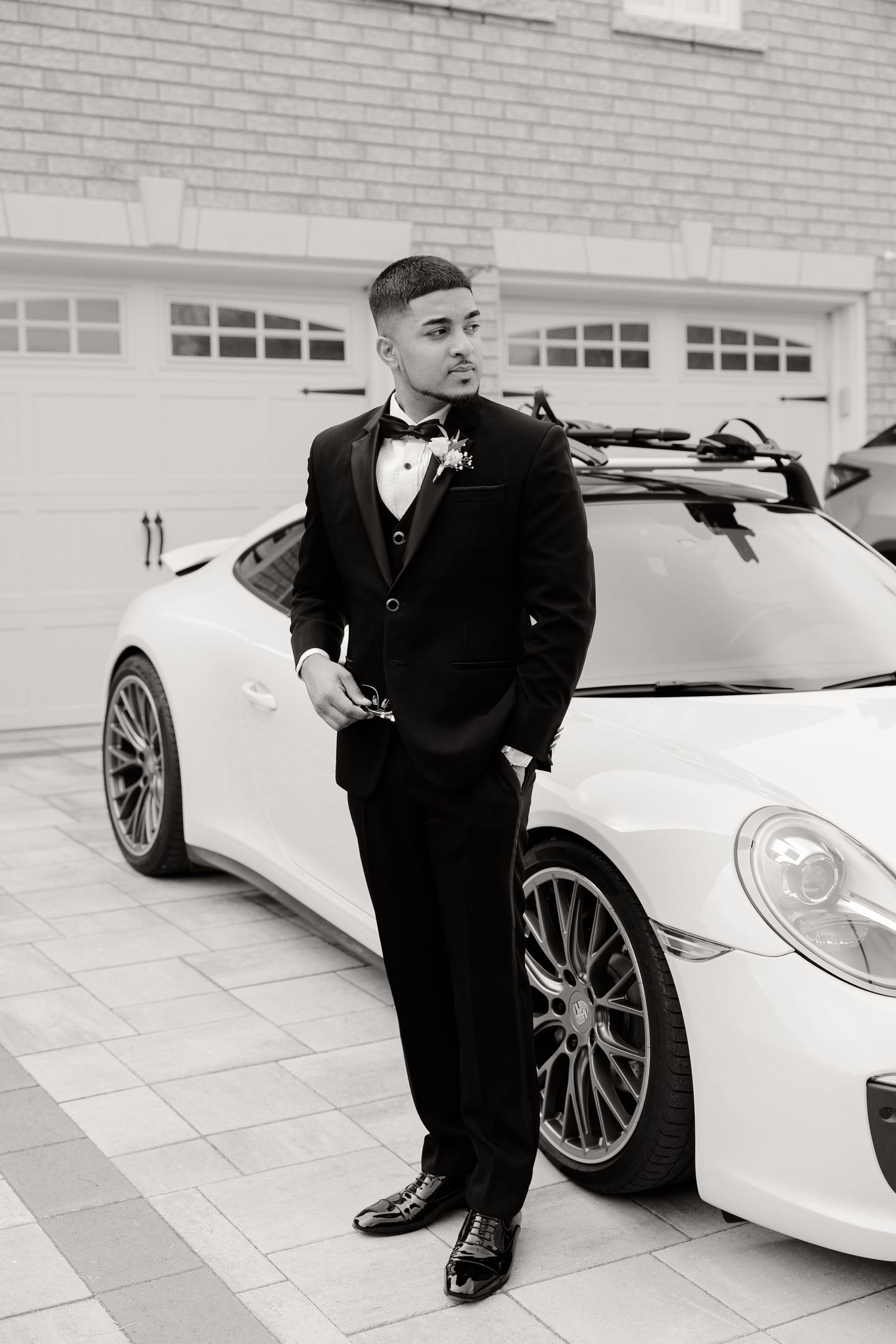 Black and white portrait of a person in formal attire posing beside a luxury Porsche sports car.