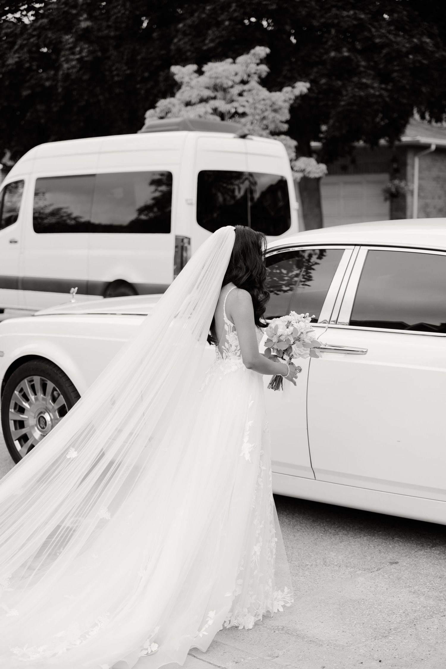 Black and white photo of bride with long veil next to luxury wedding car.