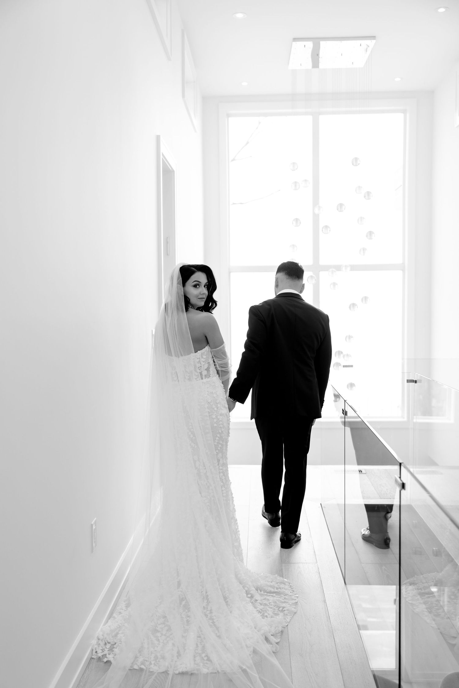Bride in lace gown glances back while walking down bright hallway with groom.