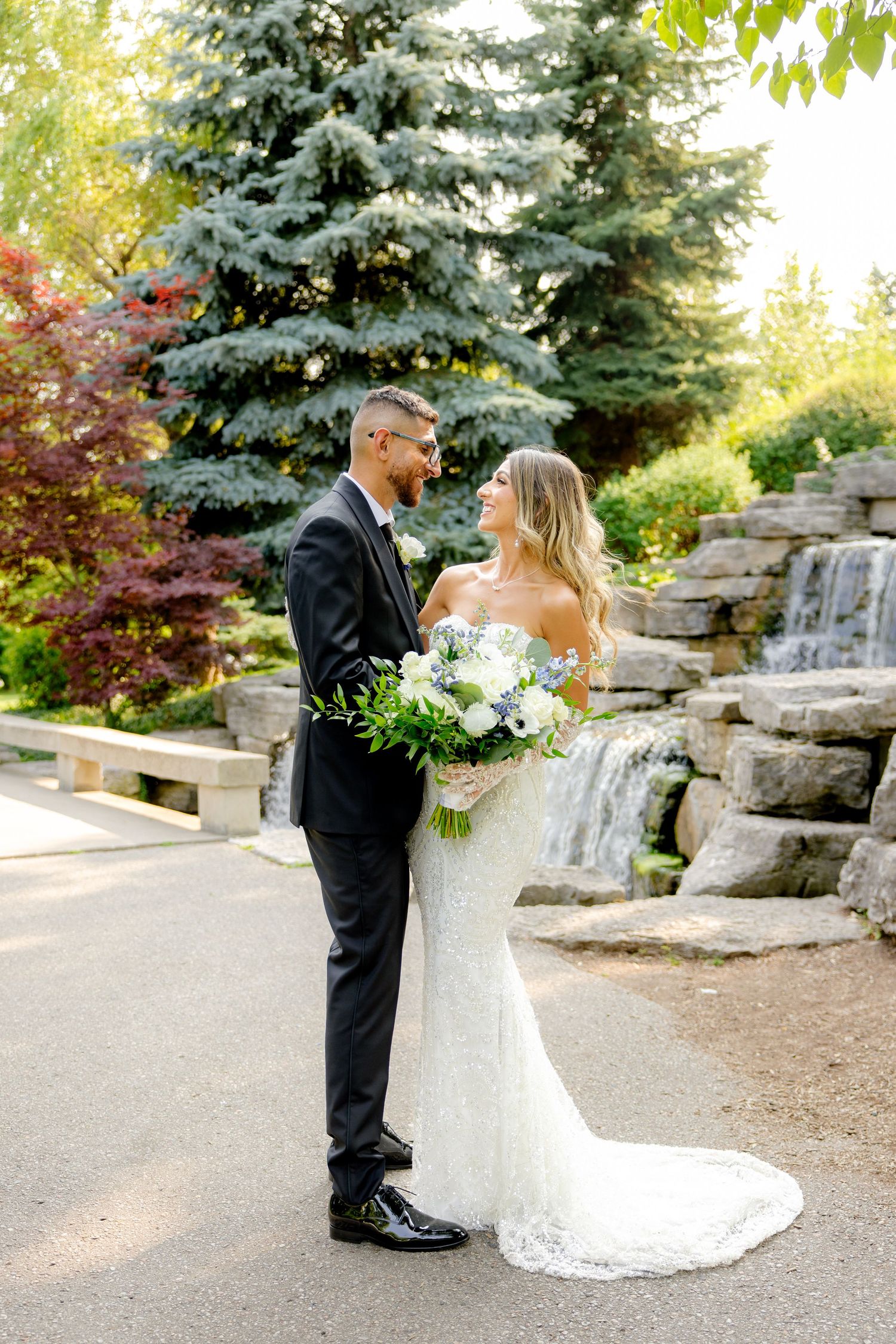 A wedding couple stands together on a garden path with a stone waterfall and evergreen trees in the background.